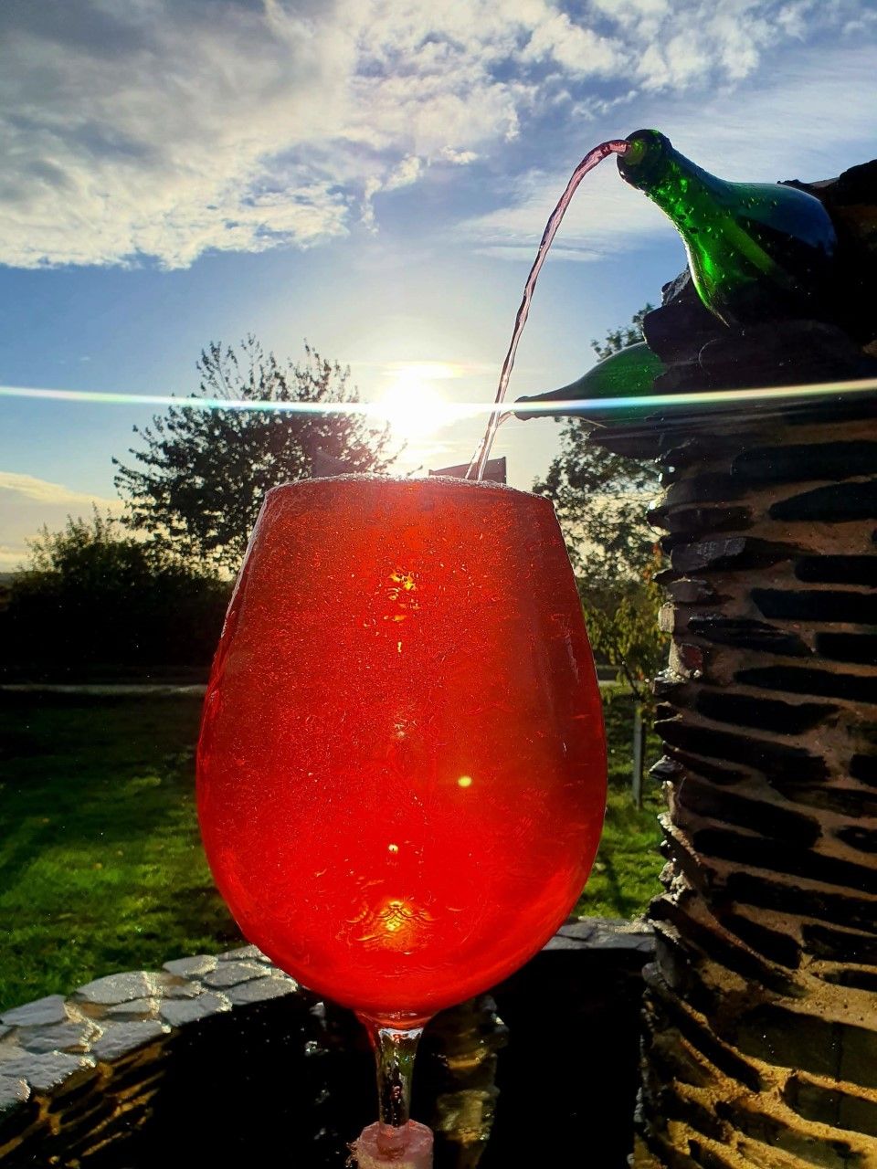 A glass of red liquid with a stream of clear liquid being poured into it, set against a backdrop of a clear sky and a structure with a stone pattern.