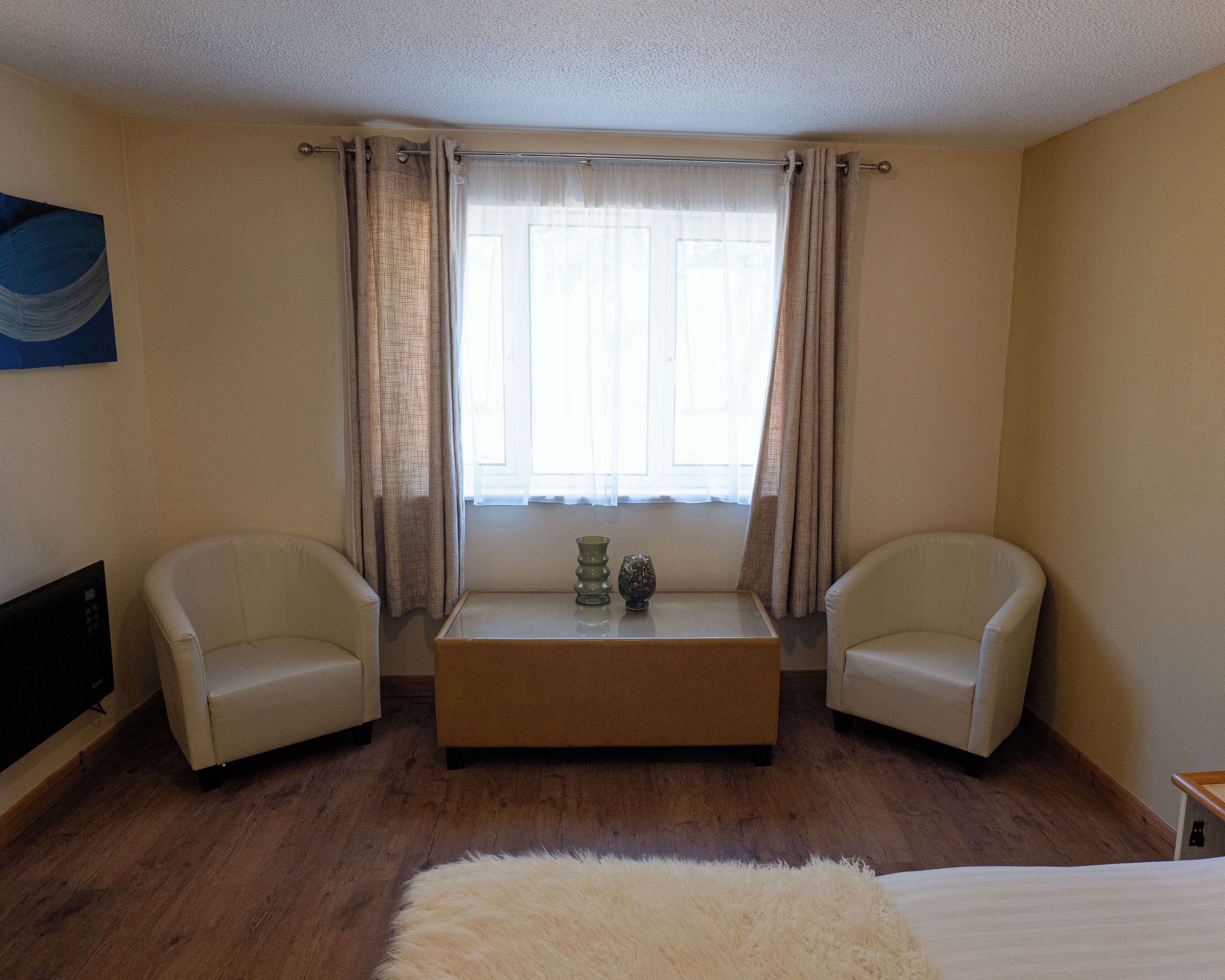 Een hotelkamer met twee fauteuils, een houten bijzettafeltje, een zacht, wit tapijt en een groot raam met gordijnen.