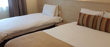 Habitación de hotel con dos camas, ropa de cama color beige, piso de madera y dos luces fijadas a la pared.