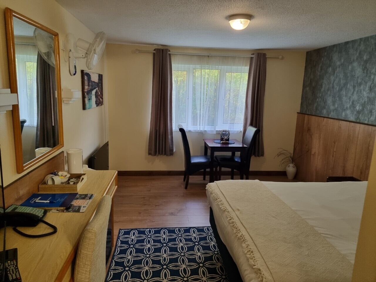 Een hotelkamer met een bed, een bureau met stoel, een klein tafeltje, een spiegel en een raam met gordijnen.