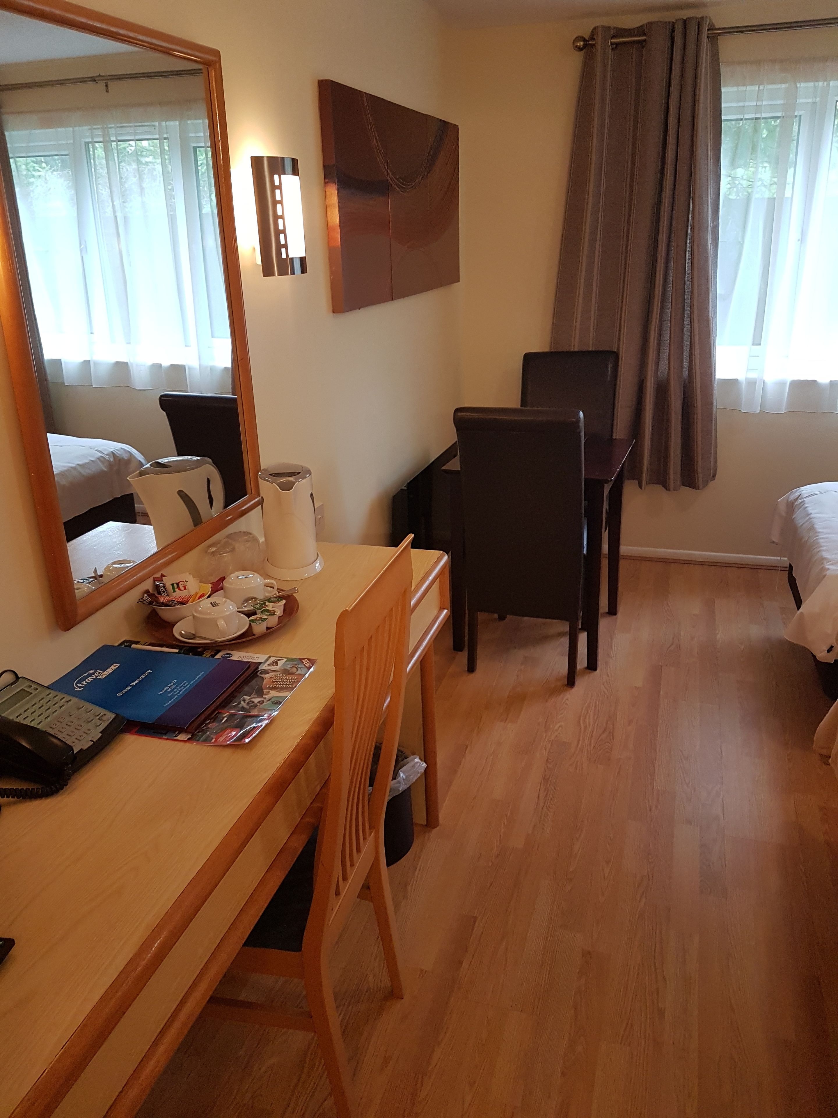 Een hotelkamer met een houten bureau, een stoel, een bed, een raam met gordijnen, een wandlamp en een spiegel.