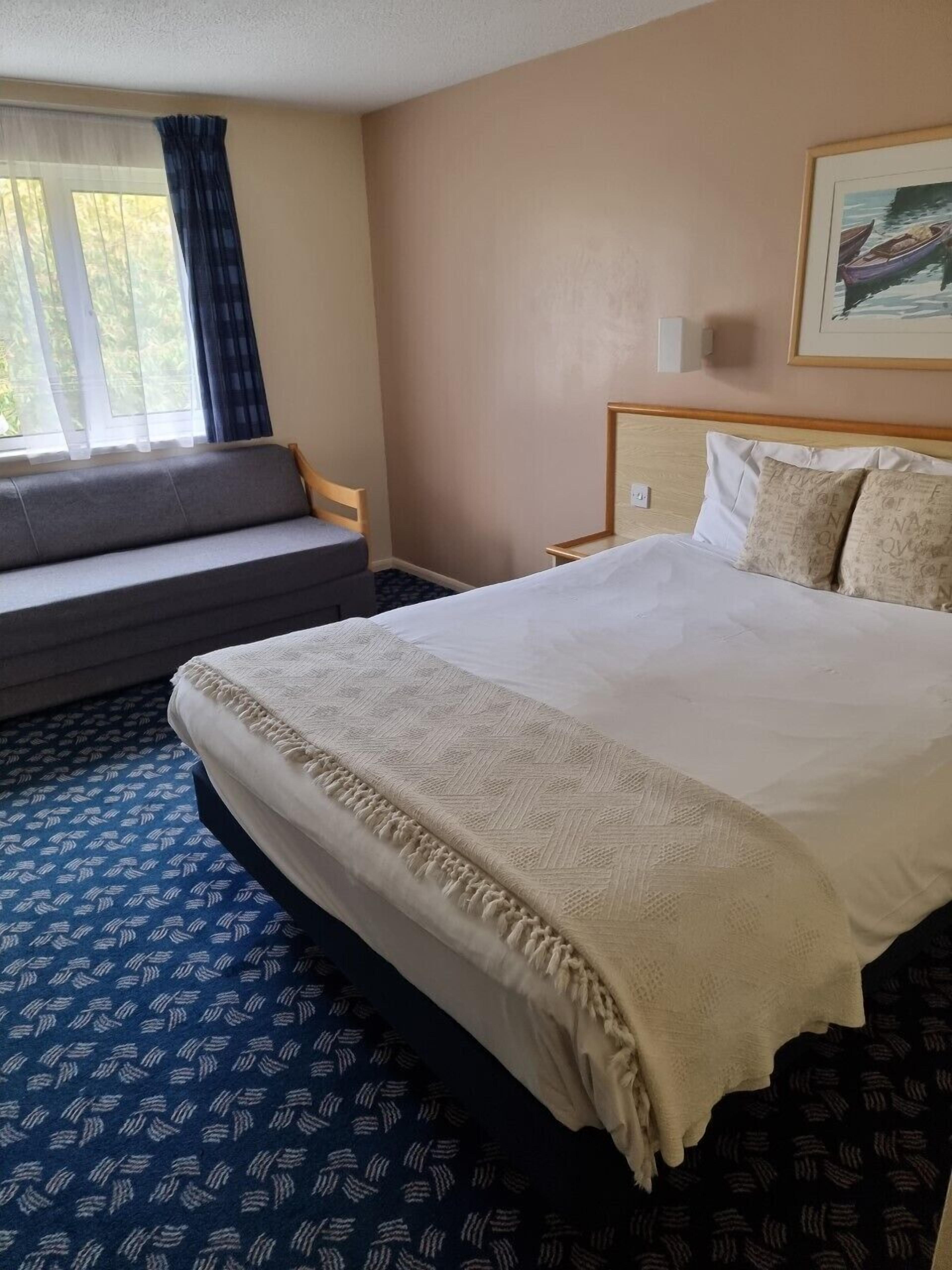 A hotel room with a bed, a sofa, a window with curtains, a painting on the wall, and a patterned carpet.