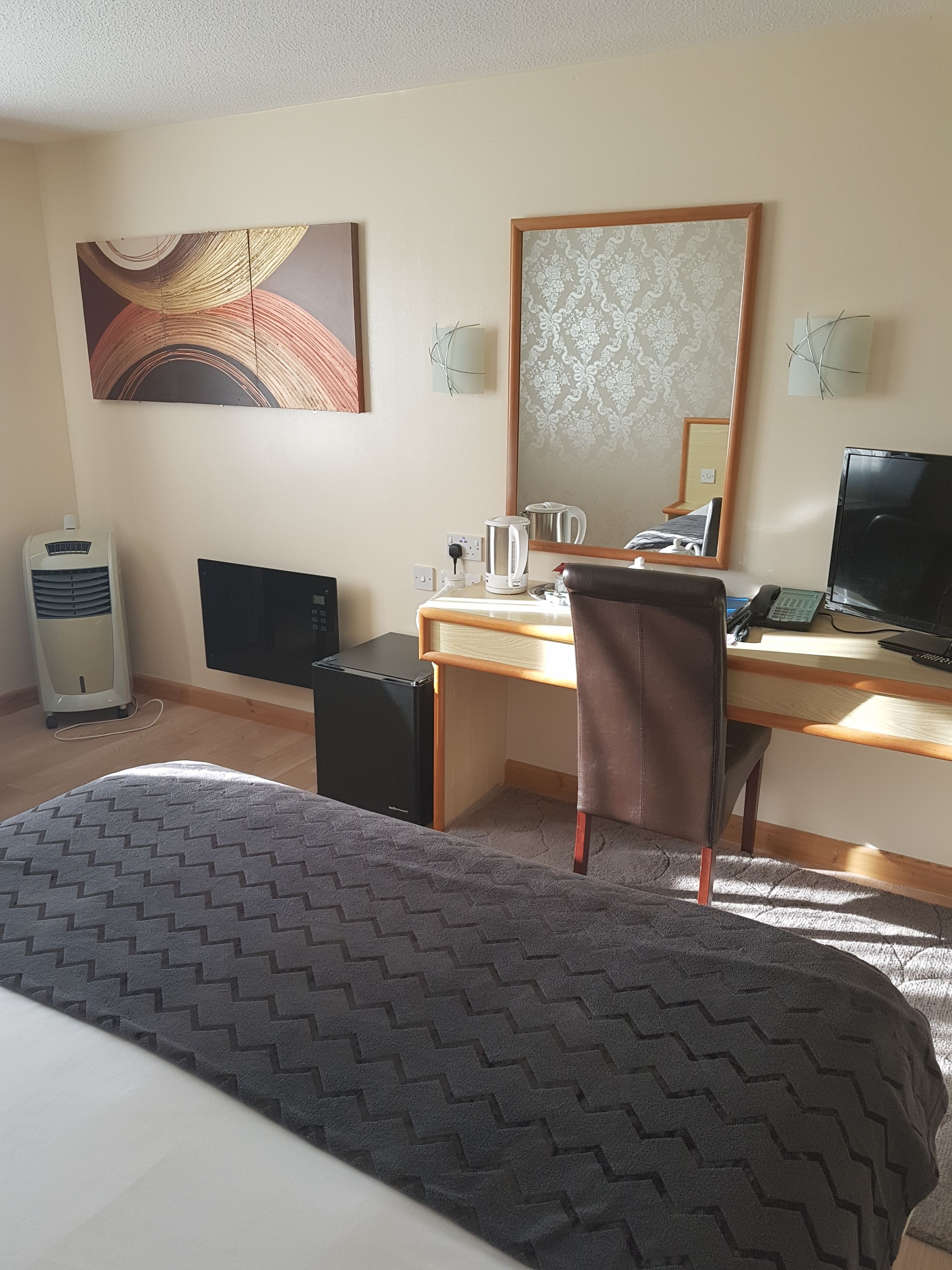 Habitación de hotel con cama, escritorio con computadora, silla, cafetera, tetera, televisión y un espejo grande.