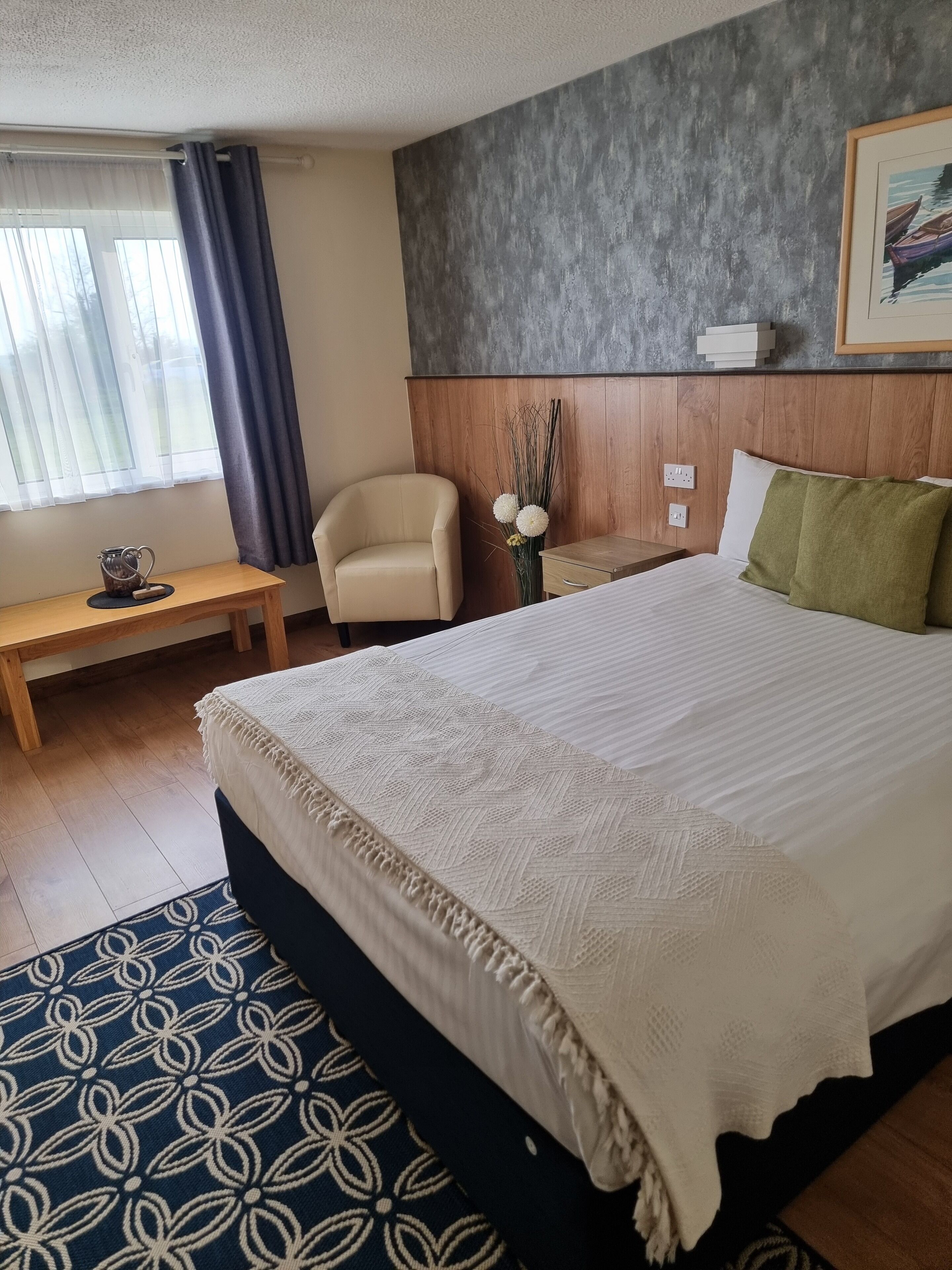 A hotel room with a large bed, a wooden nightstand, a chair, a small table, and a window with curtains.