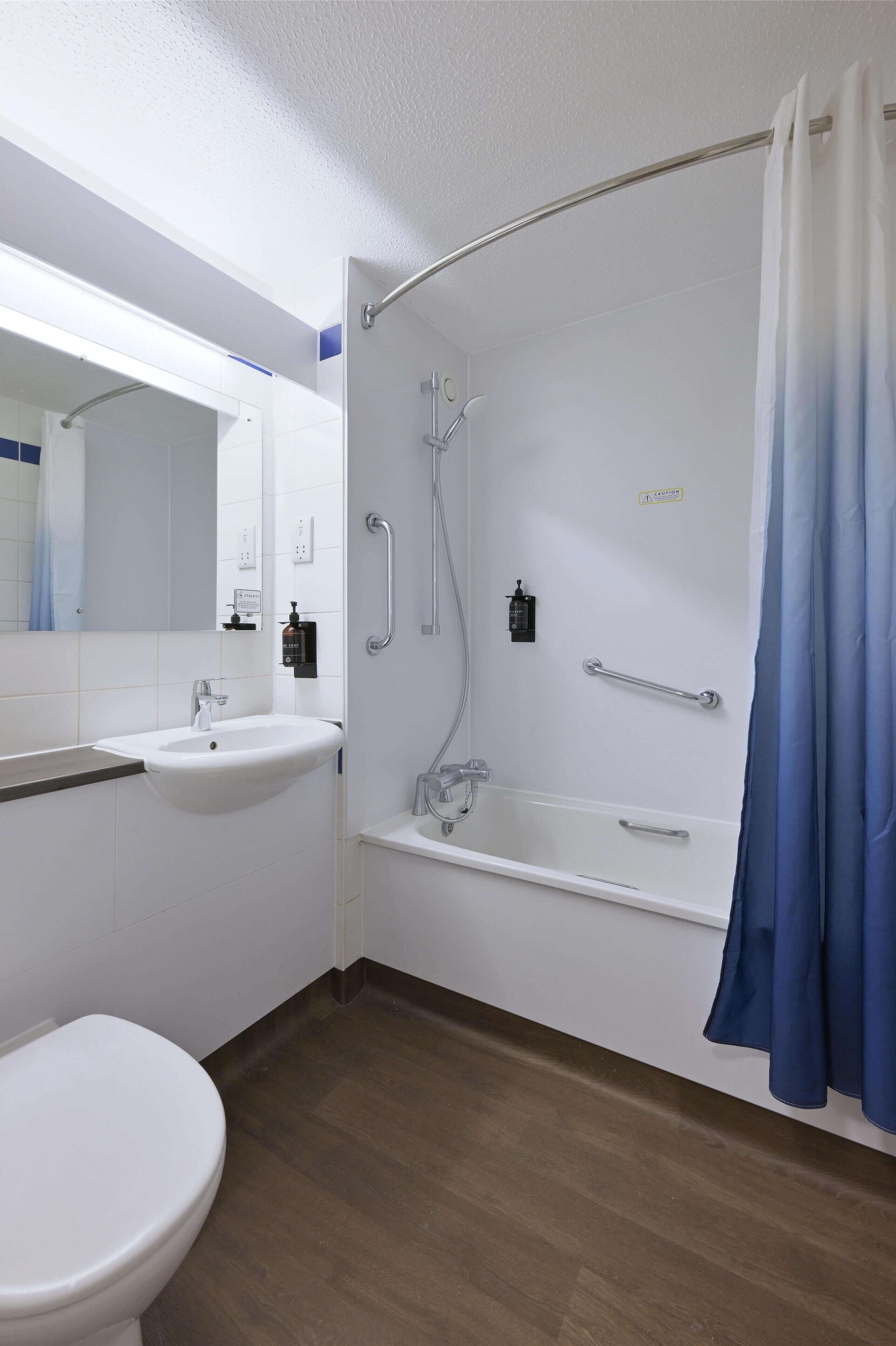 Un baño con un lavamanos blanco, un inodoro, una ducha con puerta de vidrio curva y una cortina de ducha azul.
