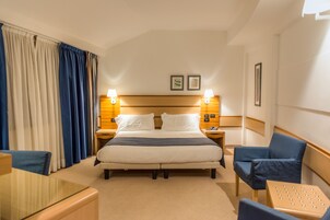 Una habitación de hotel con una cama grande, dos lámparas de noche, un cabecero, una mesita pequeña y dos sillas.