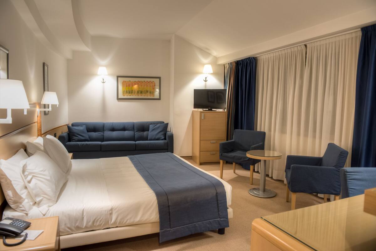 Ein Hotelzimmer mit Bett, Sofa, Schreibtisch, Stuhl, Fernseher und einem Fenster mit Vorhängen.