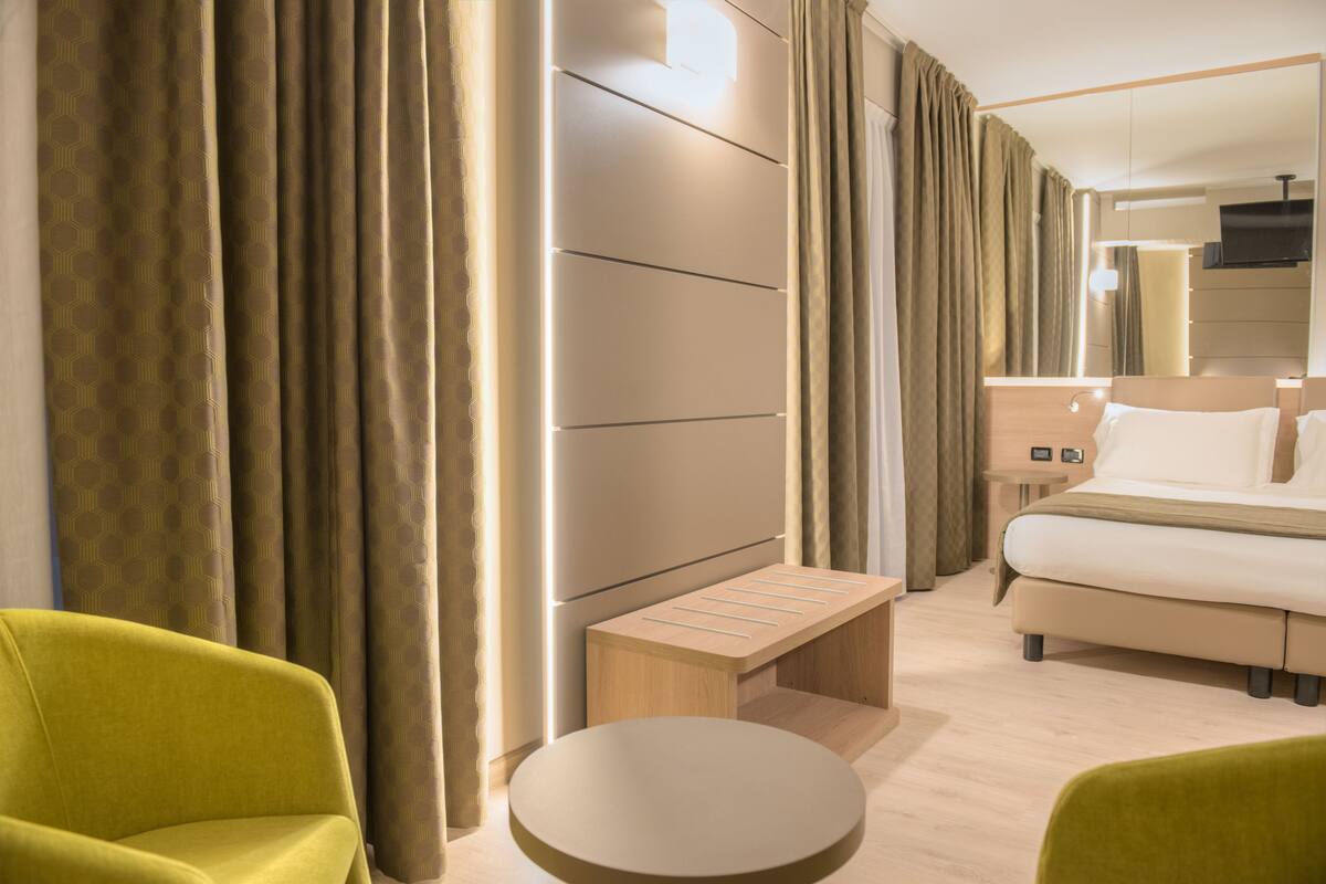 Ein Hotelzimmer mit einem Bett, einer kleinen Holzbank, einem runden Tisch und zwei grünen Stühlen.