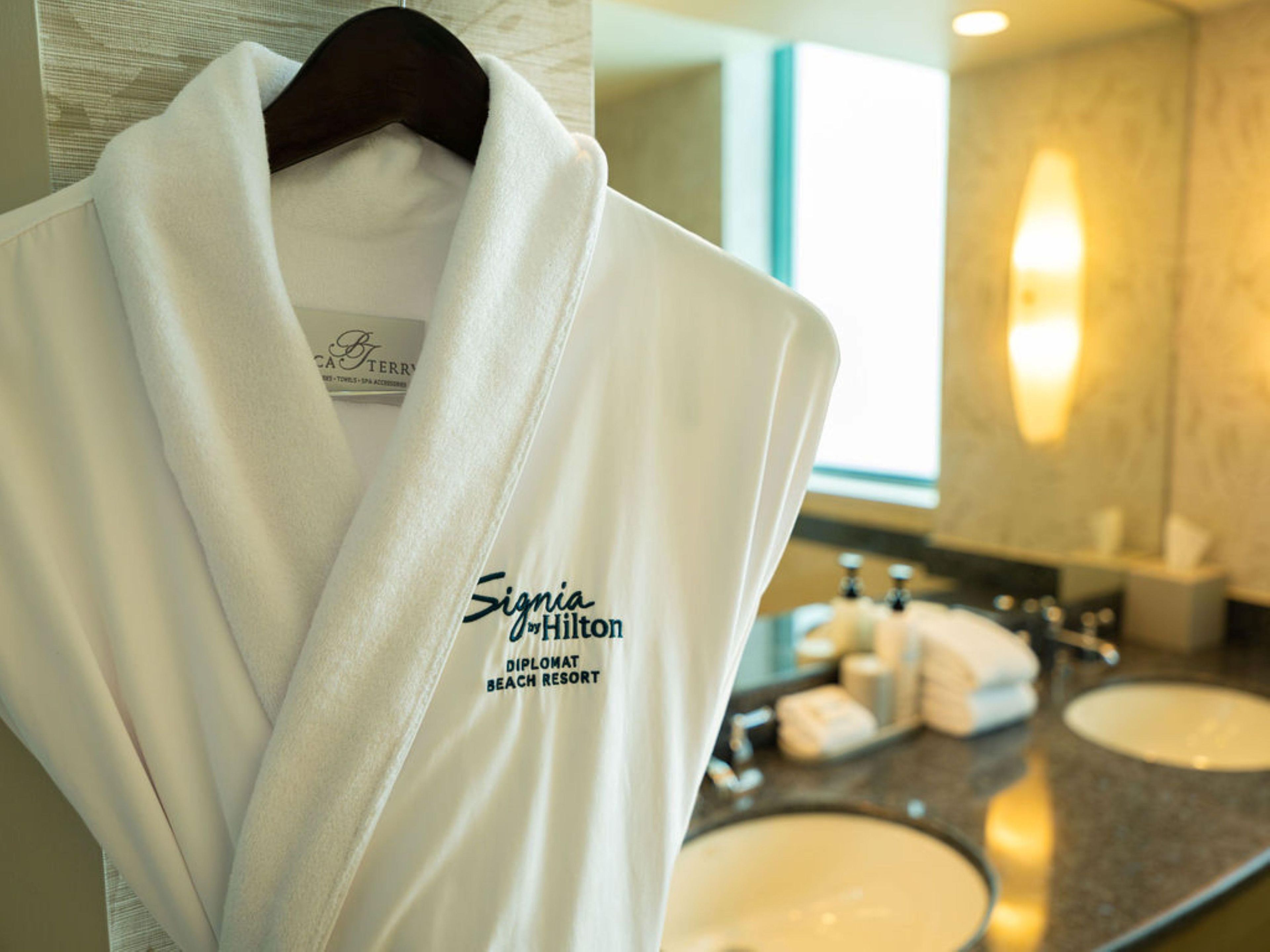A white robe with 'Signia by Hilton' and 'Diplomat Beach Resort' text, hanging on a wooden hanger in a bathroom with two sinks and a mirror.