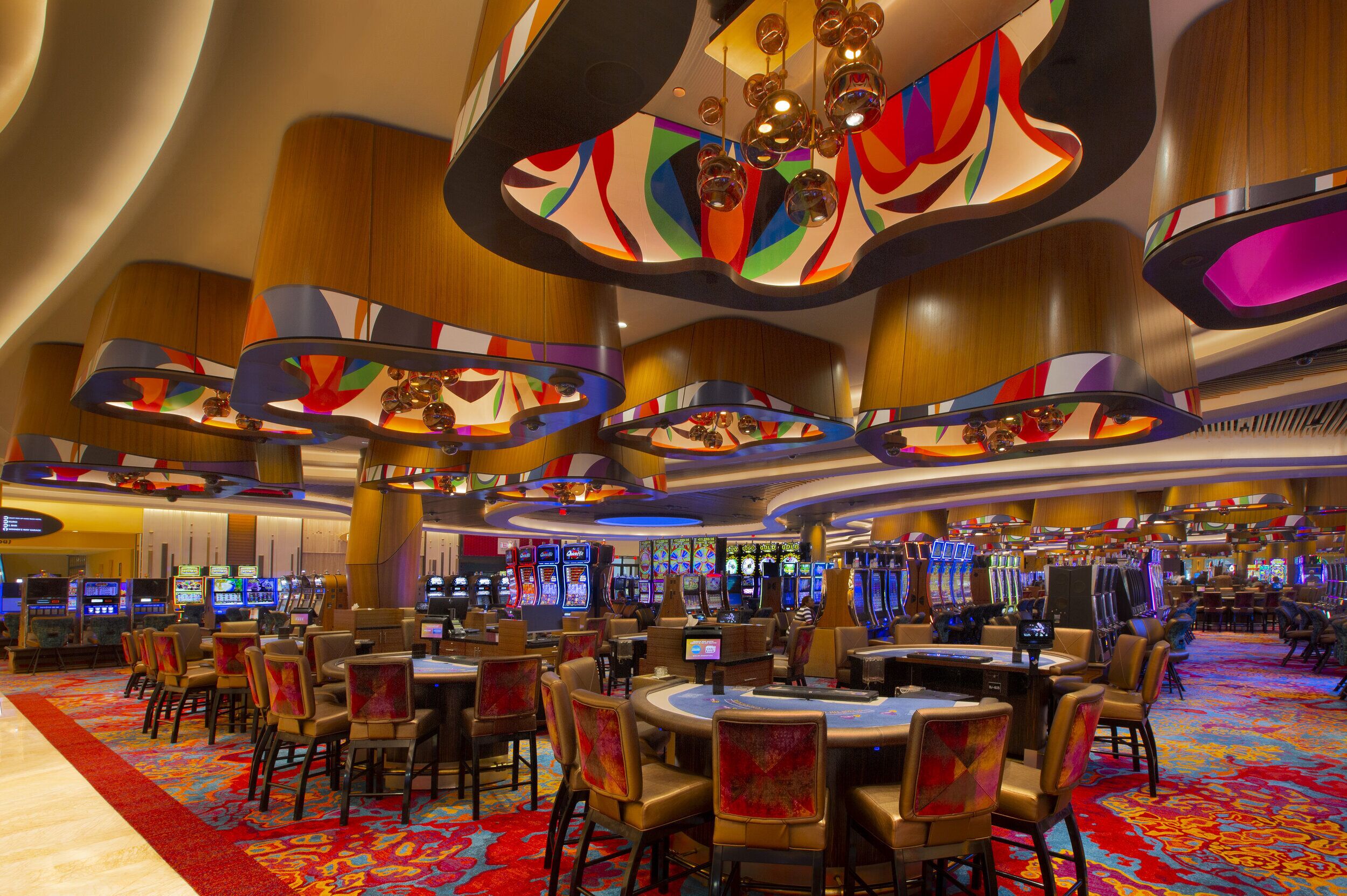 A casino with multiple gaming tables and chairs, a vibrant carpet, and a modern ceiling design.