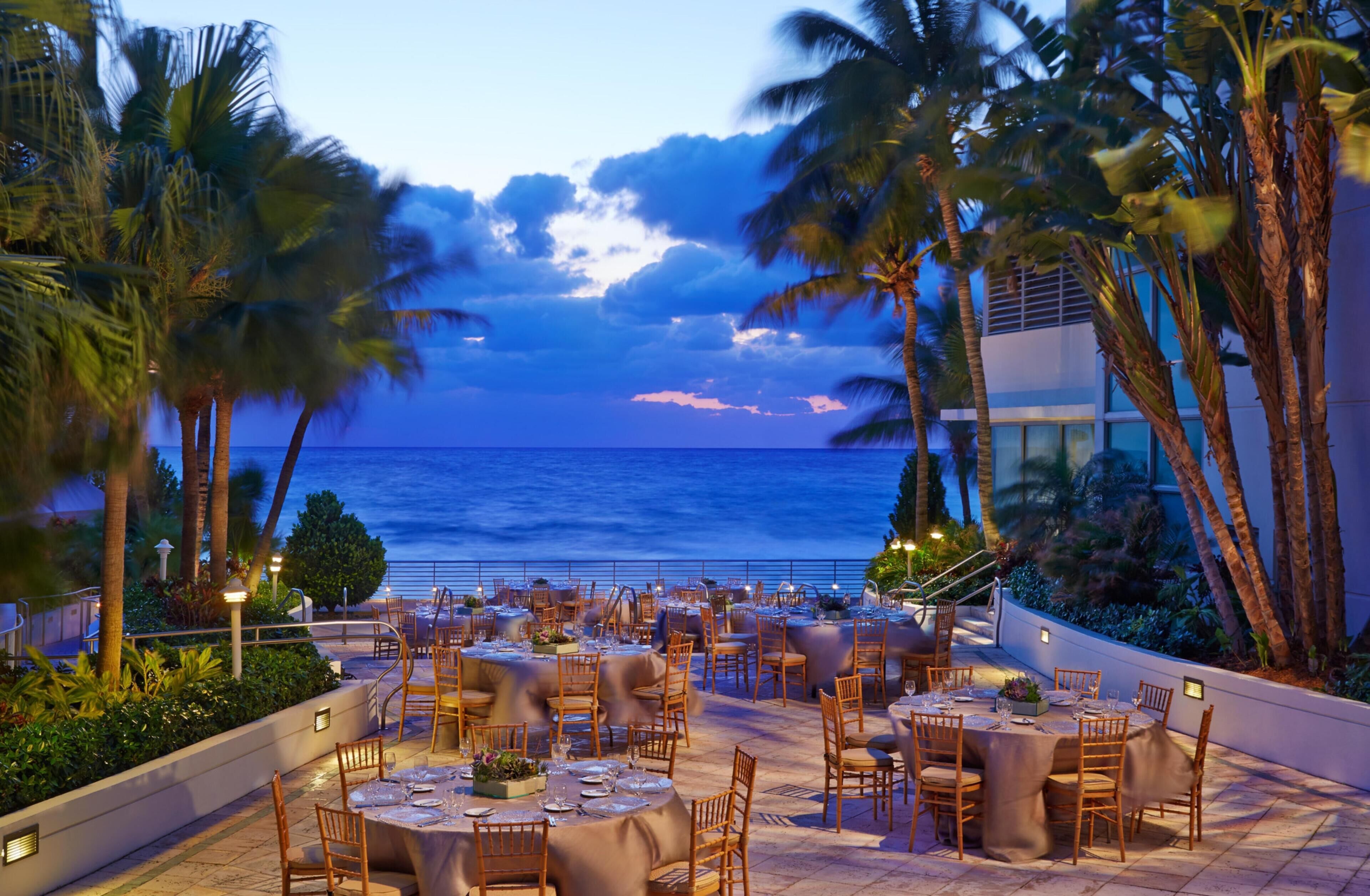 An outdoor dining area with tables and chairs, palm trees, and a view of the ocean.