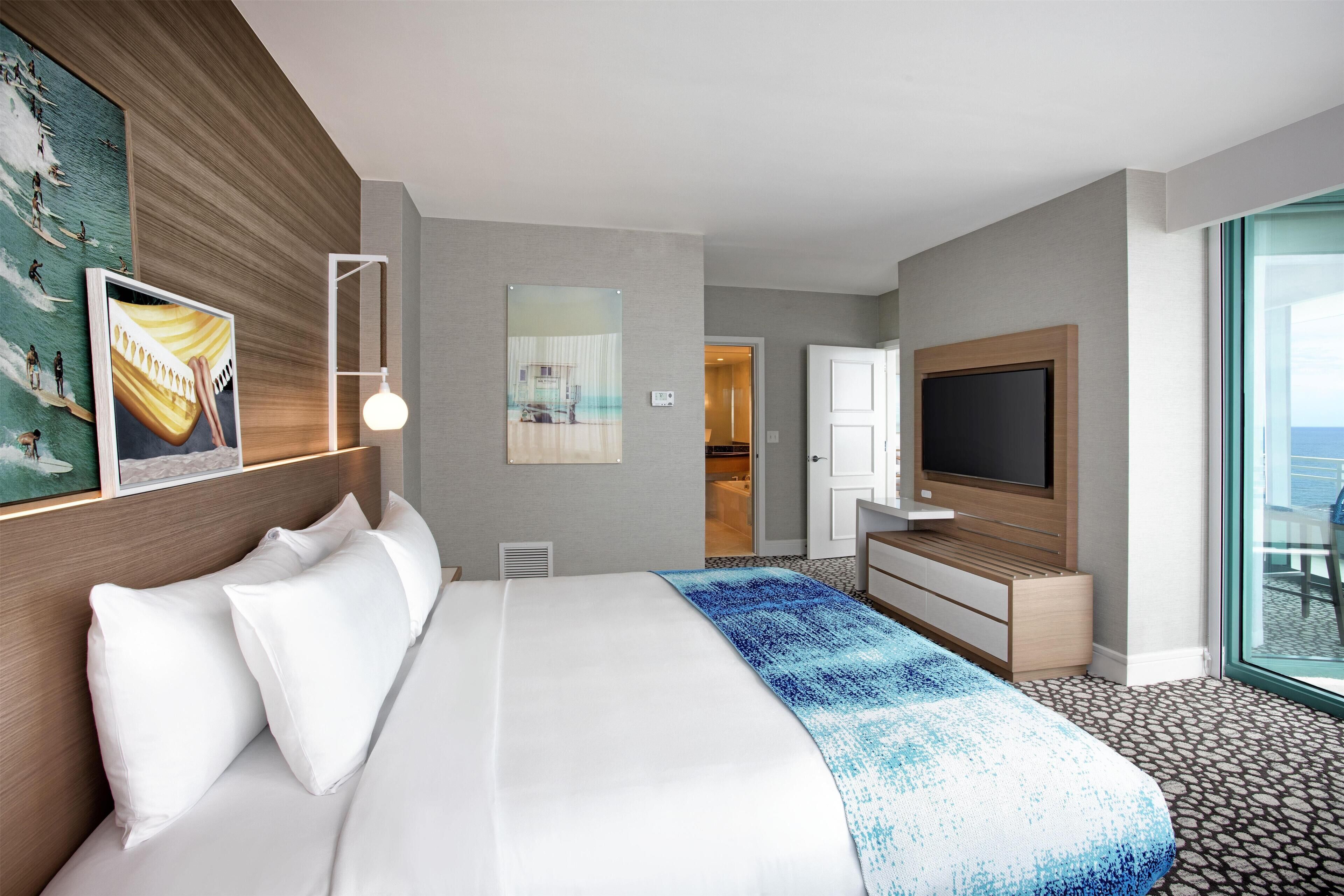 A hotel room with a bed, a TV, a large window, and a door leading to a balcony.