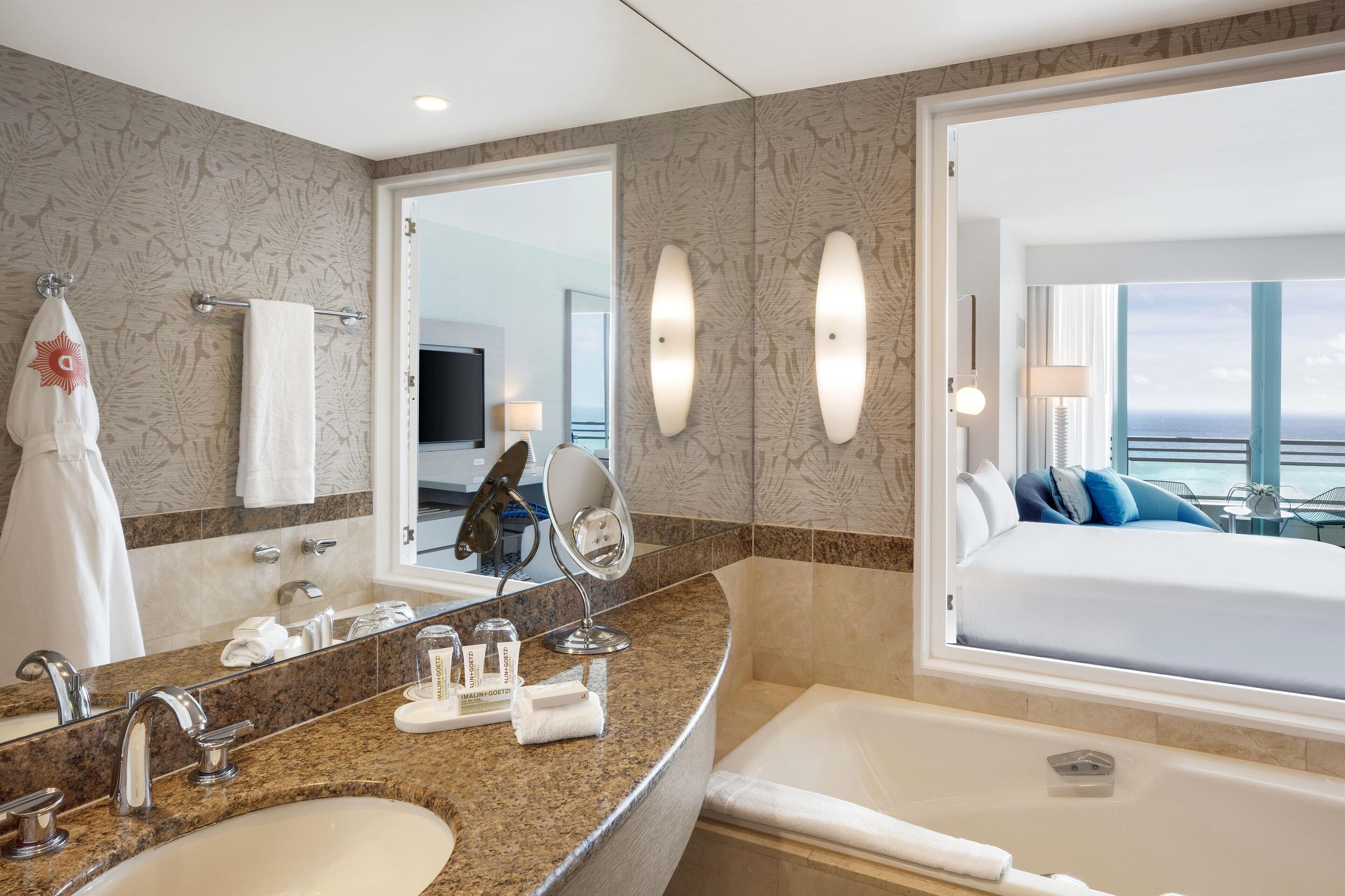 A hotel bathroom with a large mirror, two sinks, a bathtub, and a bed with a view.