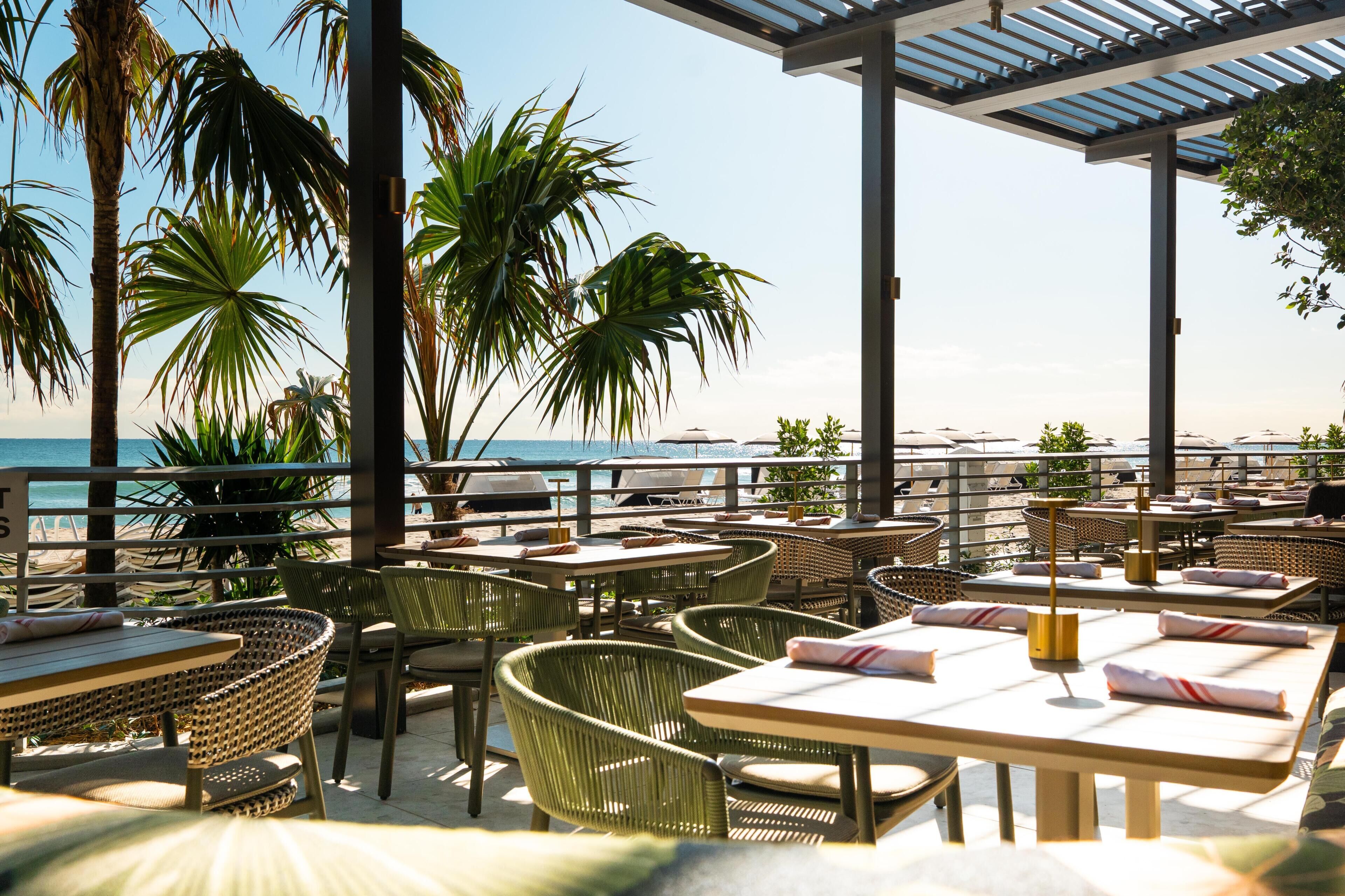 An outdoor dining area with tables and chairs, palm trees, and a view of the beach.