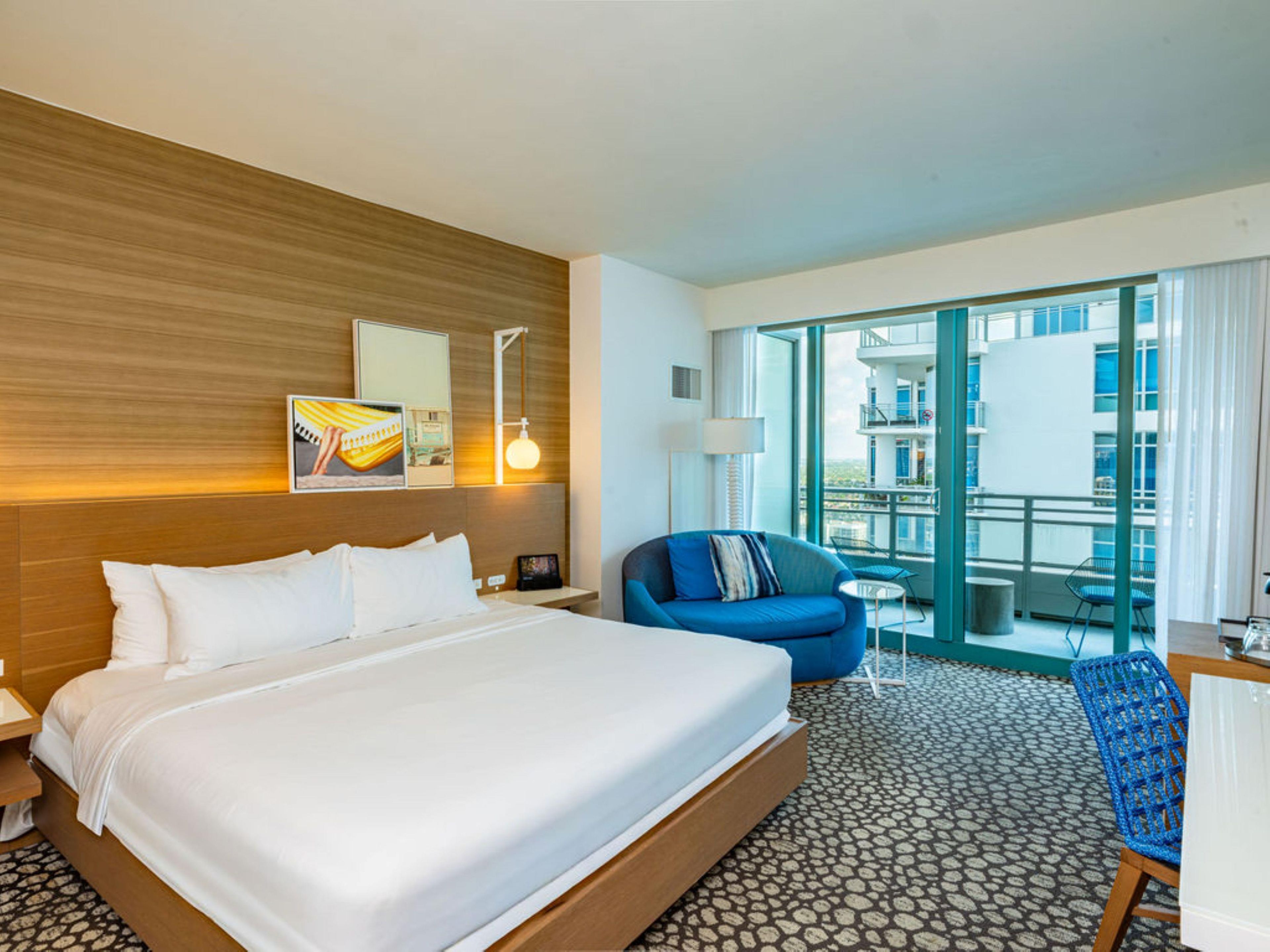 Une chambre d’hôtel avec un grand lit, un fauteuil bleu, une petite table et un balcon donnant sur la ville.