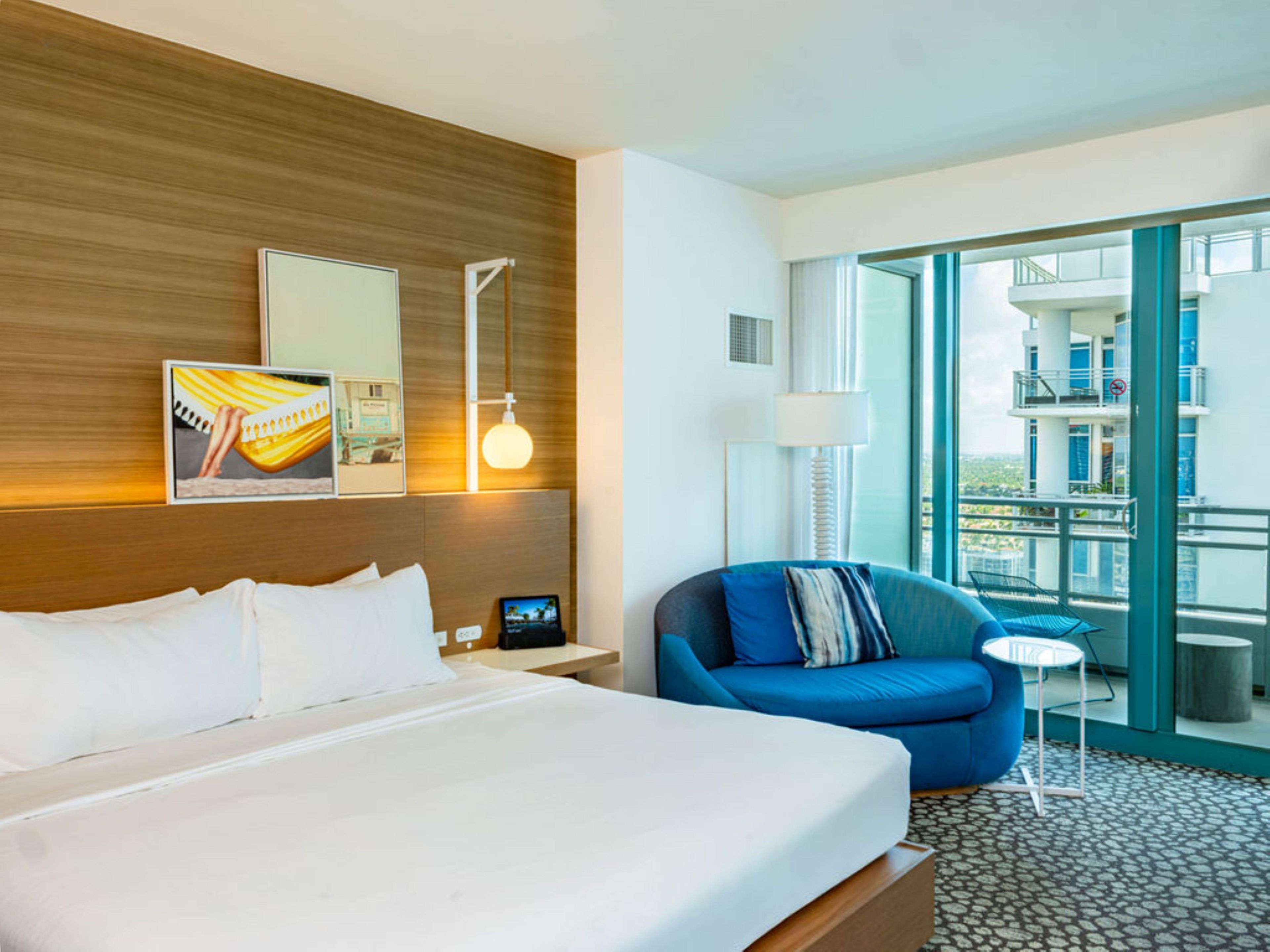 A hotel room with a large bed, a blue armchair, a small table, and a balcony with a view of the city.