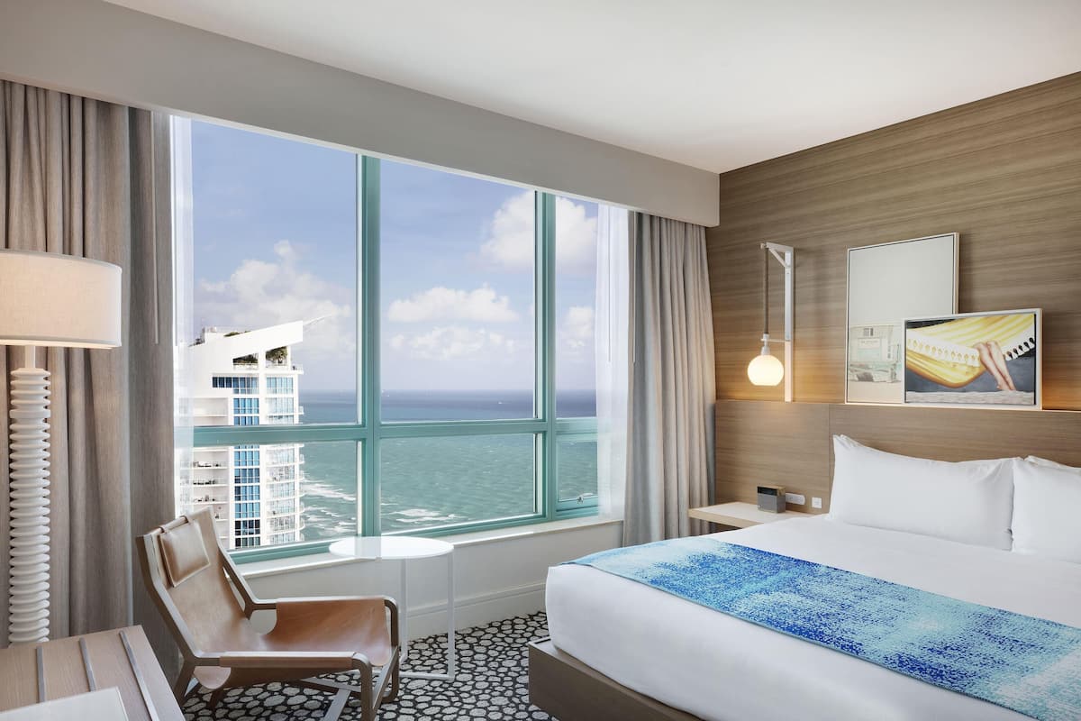 A hotel room with a large window, a bed, a chair, a desk, and a nightstand.