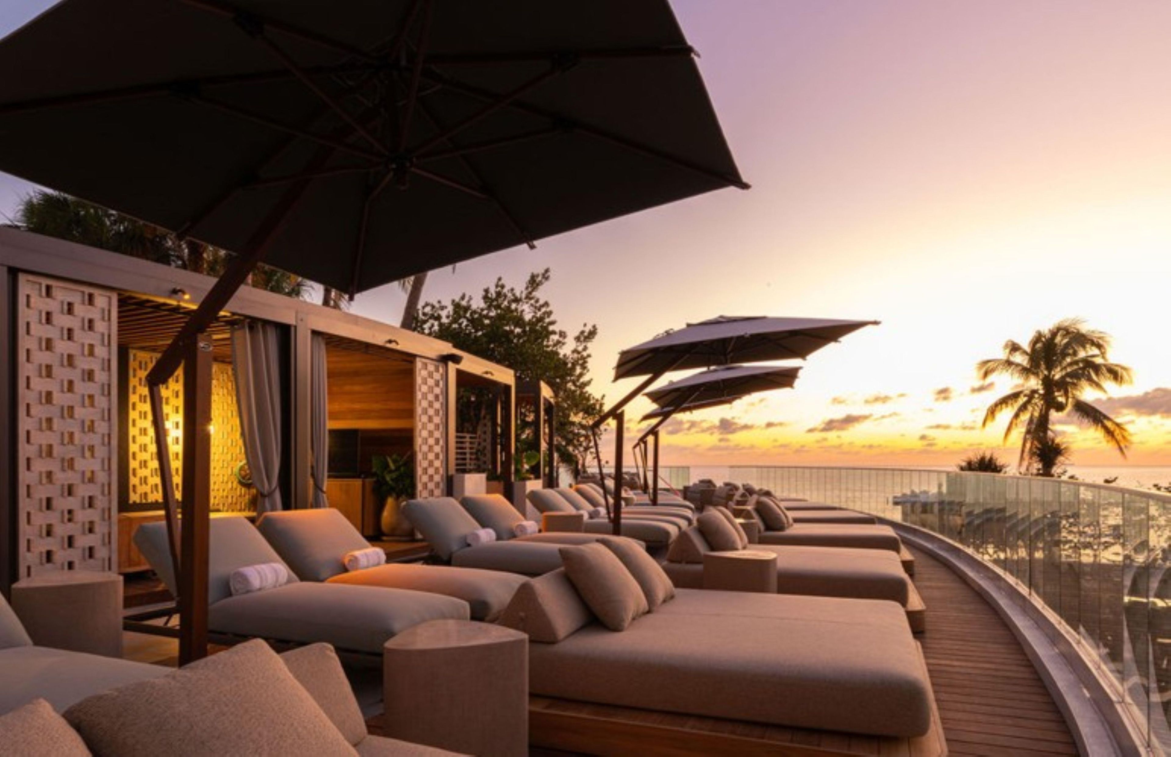 A sun lounge with multiple seating areas, umbrellas, and a view of the sunset over the ocean.
