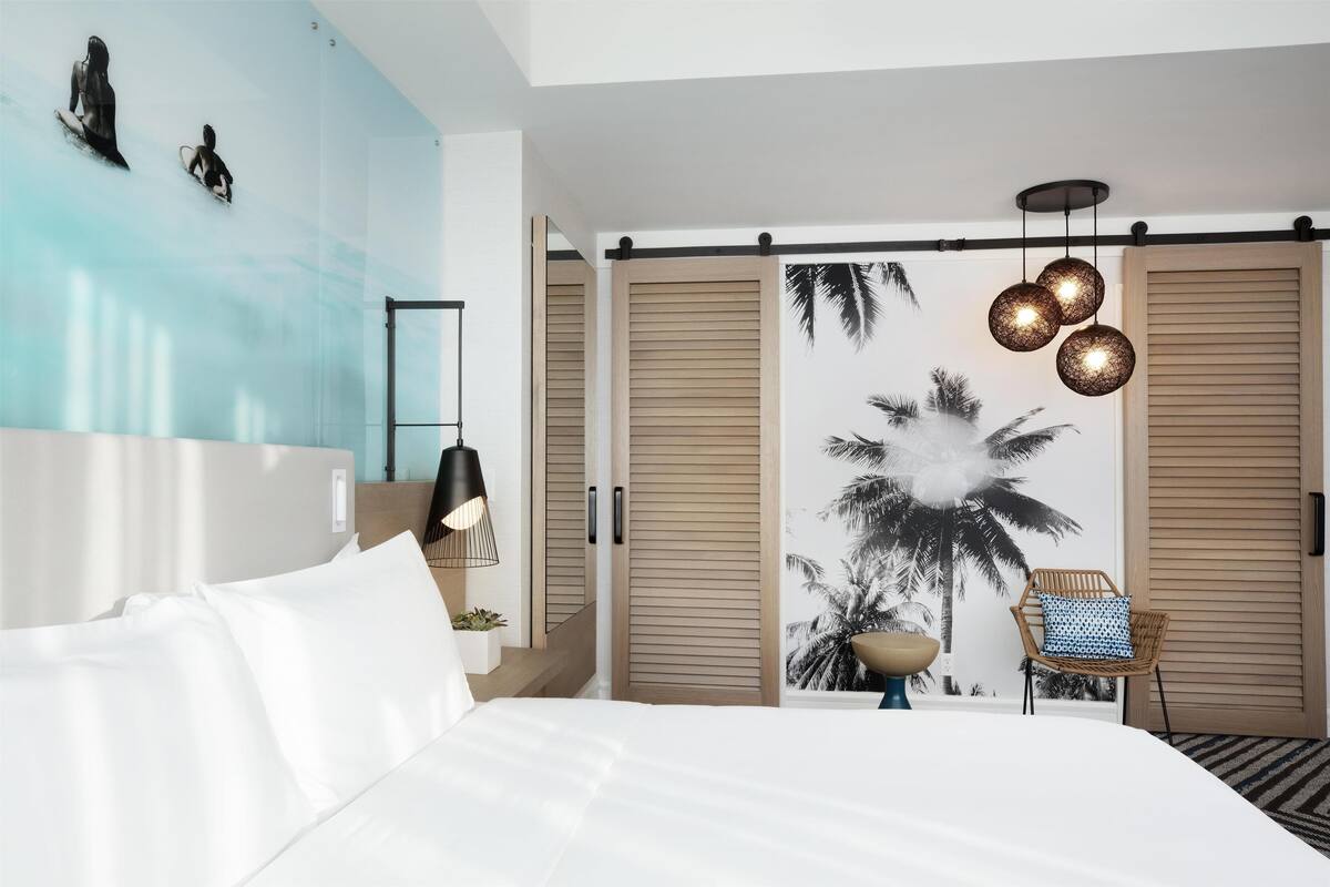 A bedroom with a bed, a chair, a wall with a palm tree mural, and a sliding door.