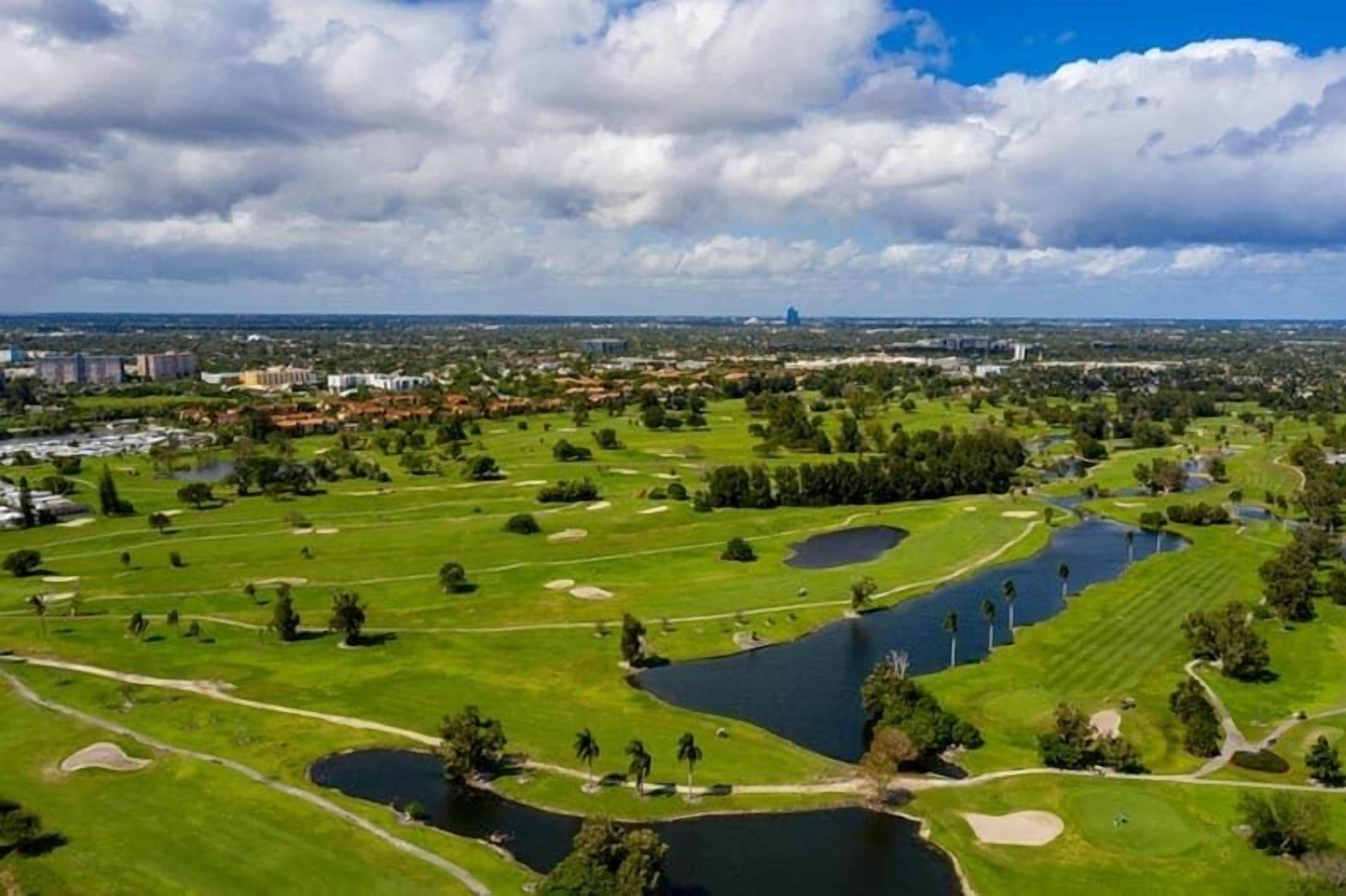 A golf course with multiple ponds, surrounded by greenery and urban buildings in the background.