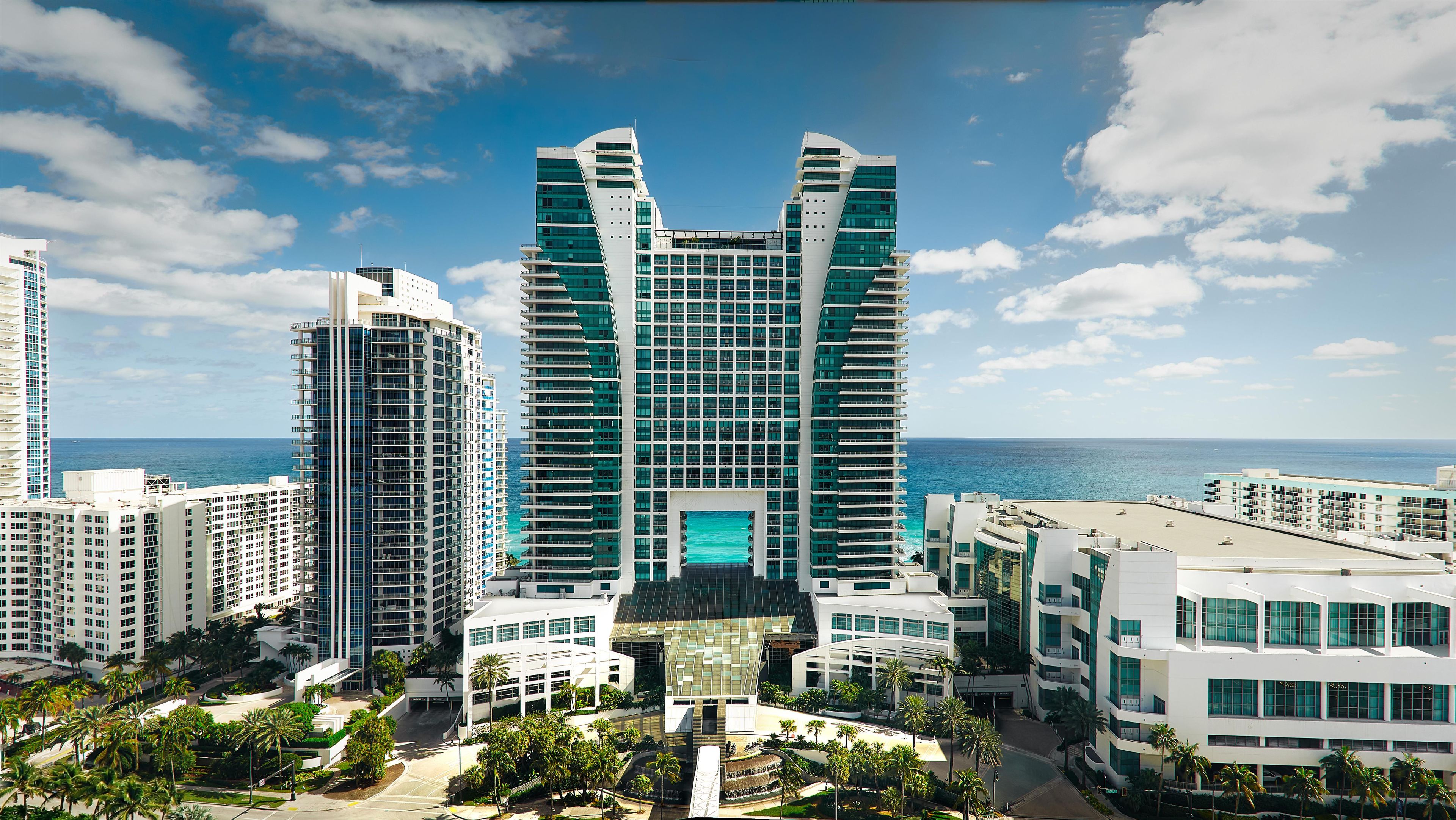 A modern high-rise hotel with a central atrium, surrounded by palm trees and a view of the ocean.
