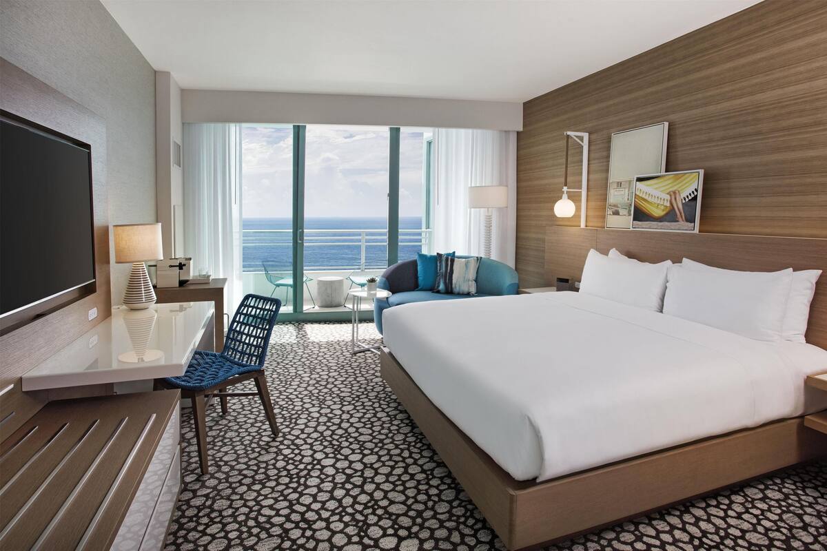 A hotel room with a large bed, a desk, a chair, a TV, and a view of the ocean.