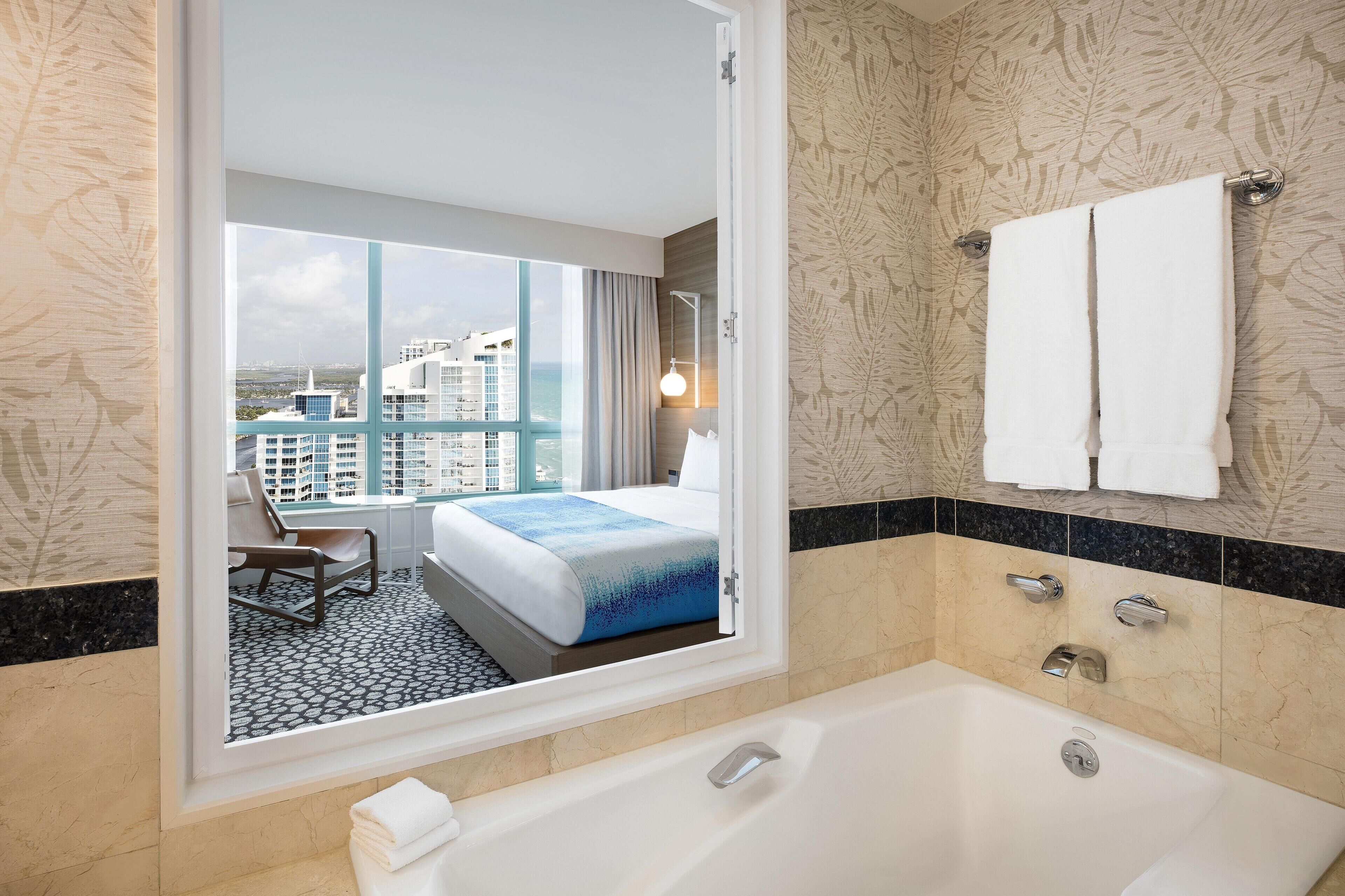 A modern hotel room with a large bathtub, a balcony with a view of the city, and a neatly made bed.