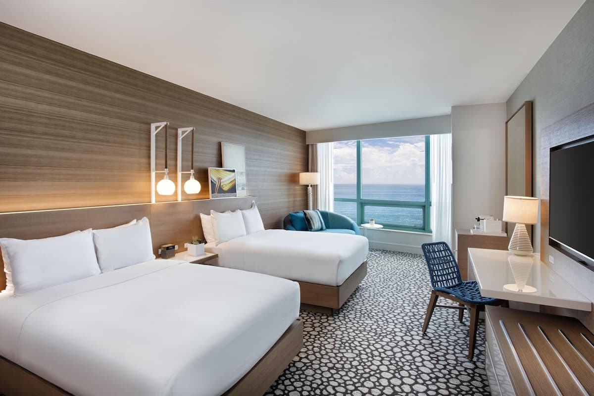 A hotel room with two beds, a desk, a chair, a TV, and a large window with a view of the ocean.