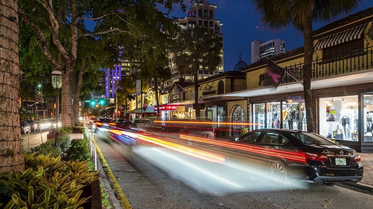 A nighttime street view with a car driving past, illuminated storefronts, and a building with a balcony.