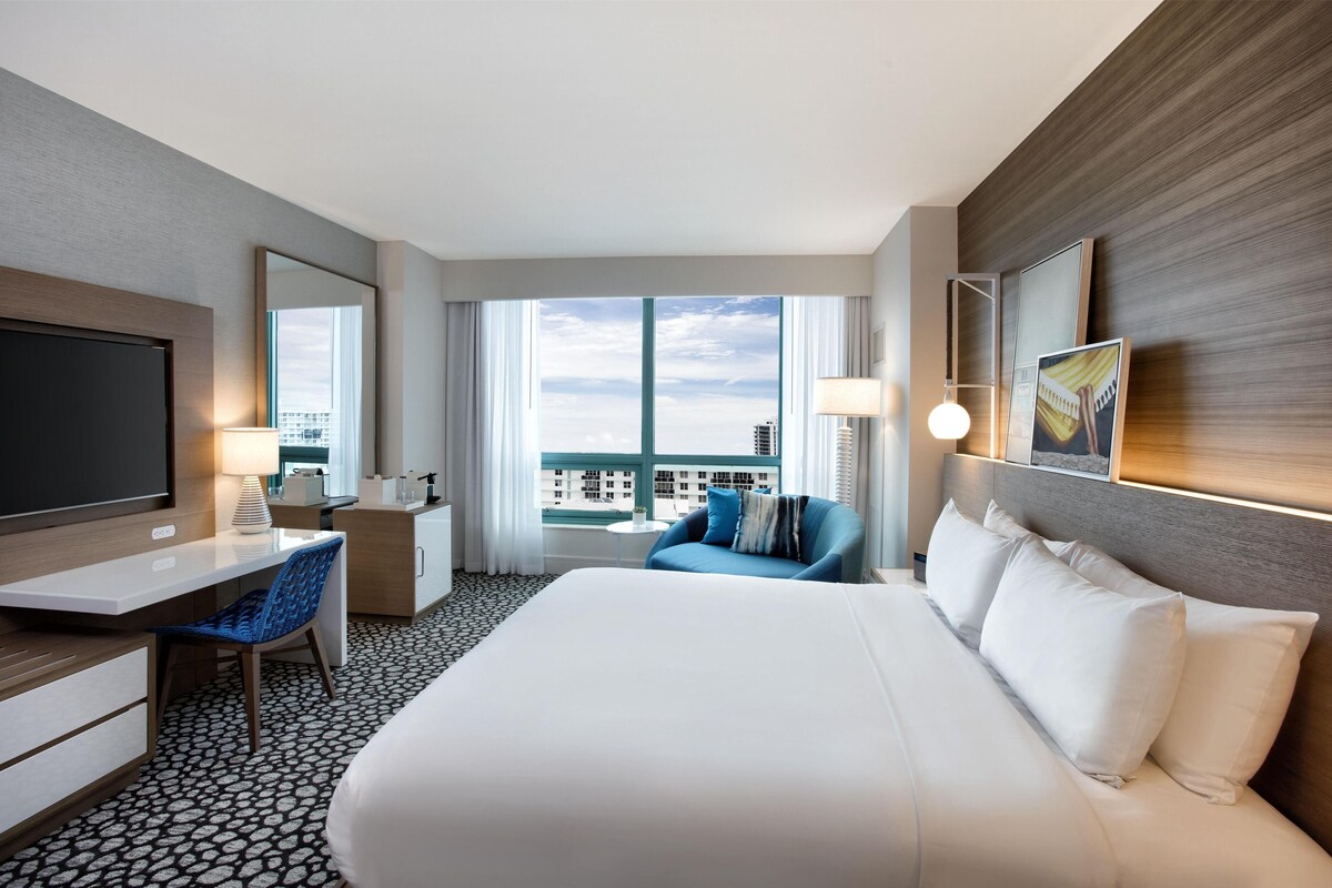 A hotel room with a large bed, a desk, a chair, a TV, a lamp, and a view of the city.