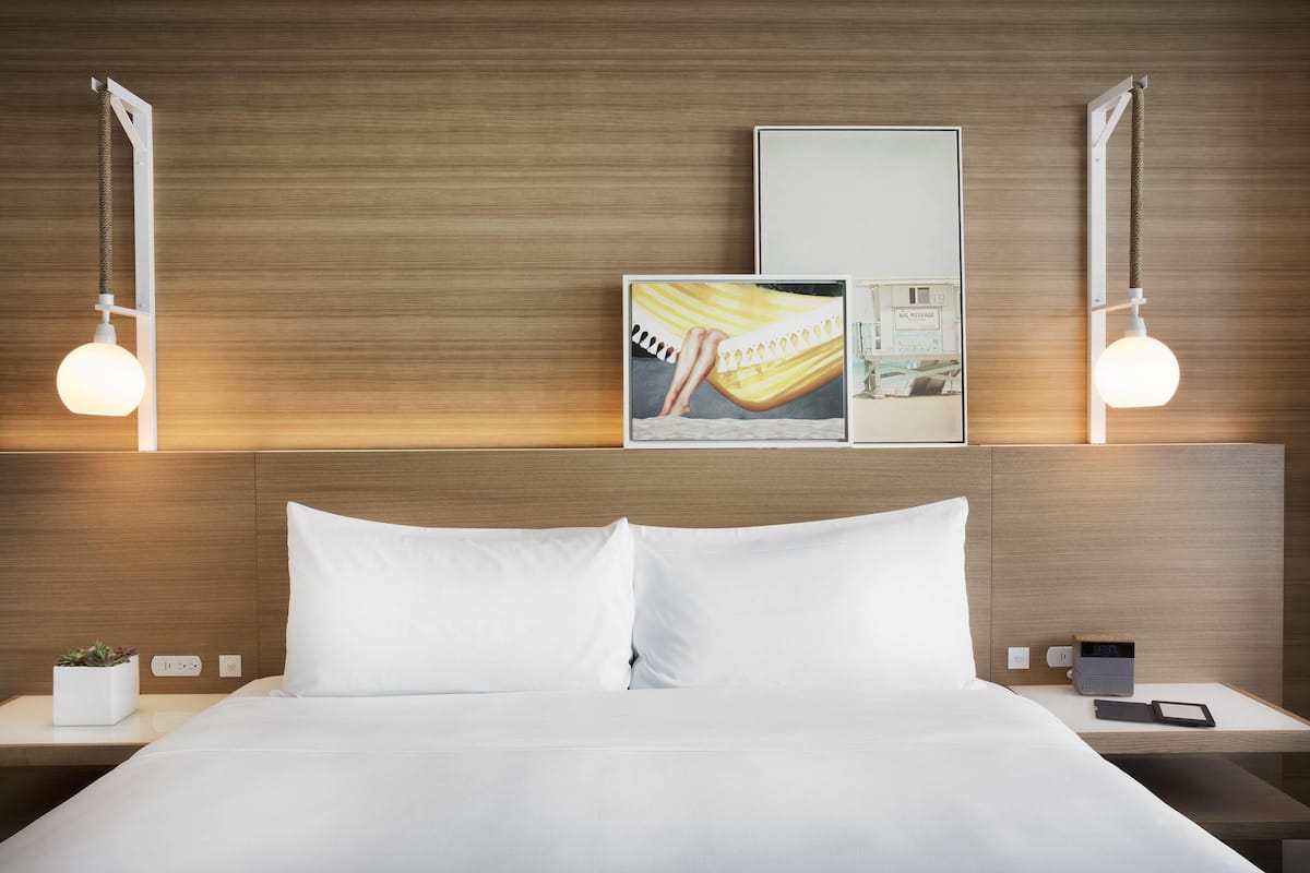 A hotel room with a bed, two bedside tables, a nightstand, a wall-mounted light fixture, a framed picture, and a digital clock.