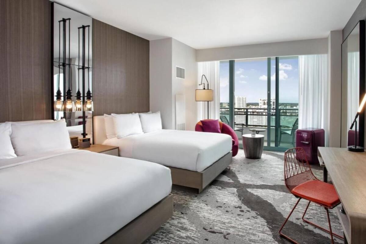 A hotel room with two beds, a desk, a chair, a lamp, and a view of the city.