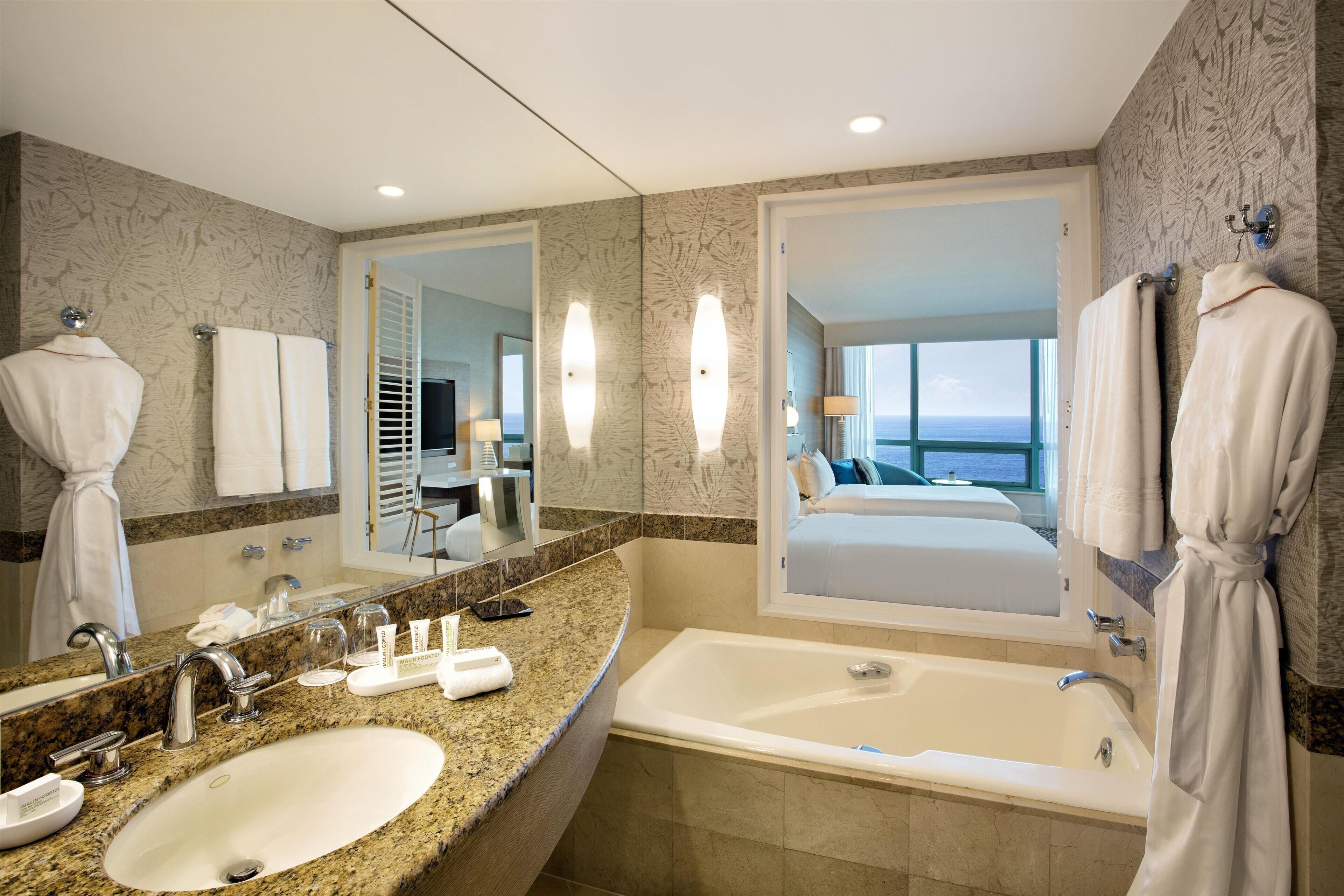 A hotel bathroom with a large sink, a bathtub, a mirror, and a view of a bedroom.