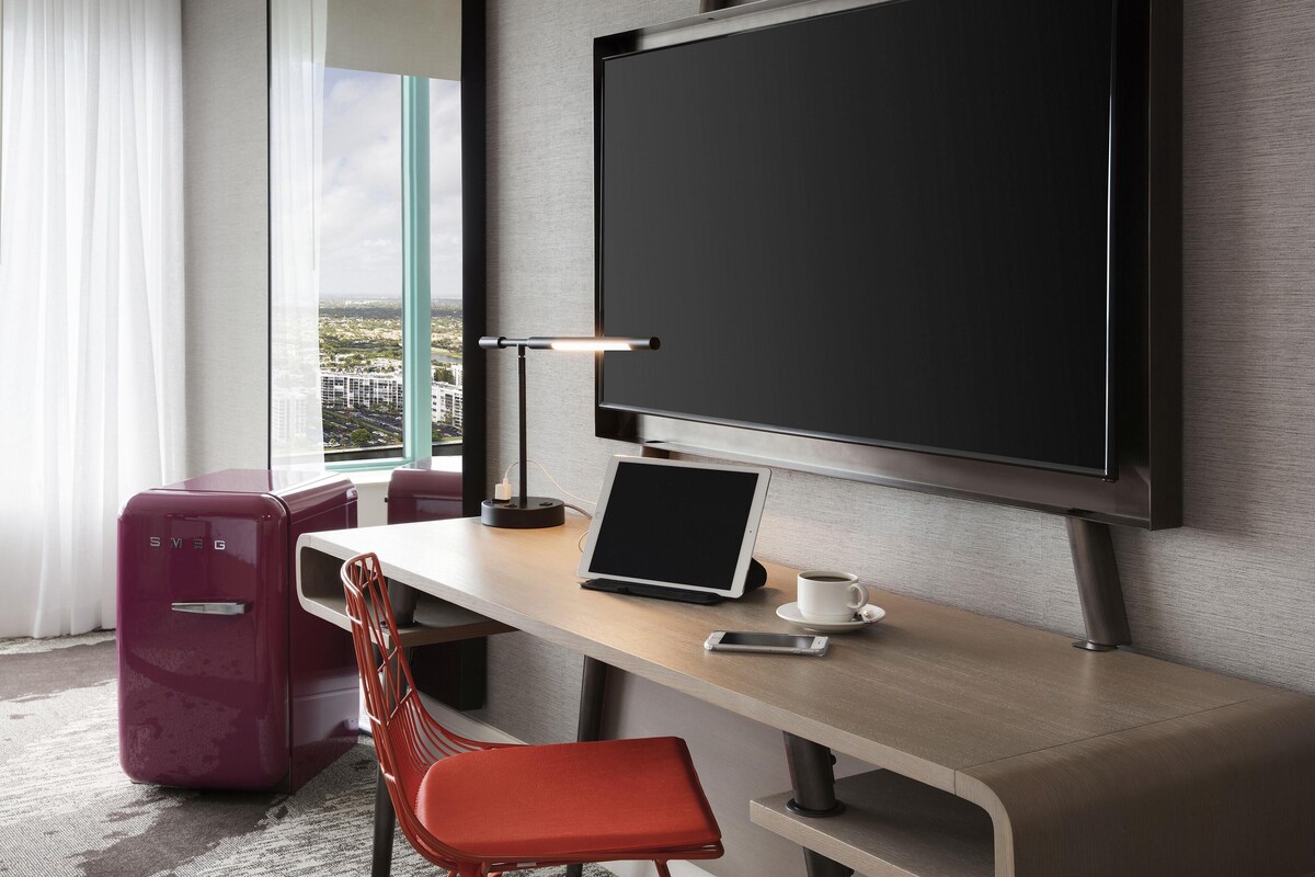 A modern hotel room with a large window, a desk with a laptop, a flat-screen TV, a red chair, and a suitcase.