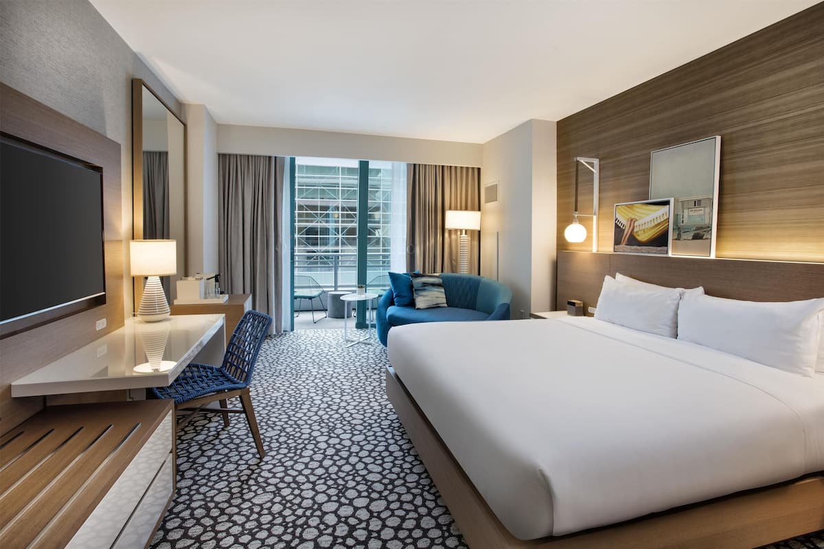 A hotel room with a large bed, a desk, a chair, a TV, a lamp, and a view of the city through the window.