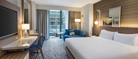 A hotel room with a large bed, a desk, a chair, a TV, a lamp, and a view of the city through the window.