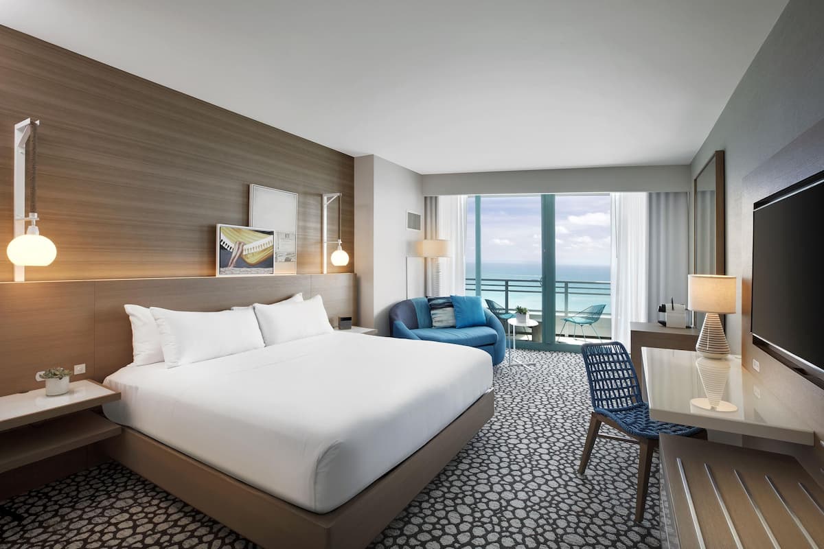 A hotel room with a large bed, a desk with a TV, a chair, and a view of the ocean.