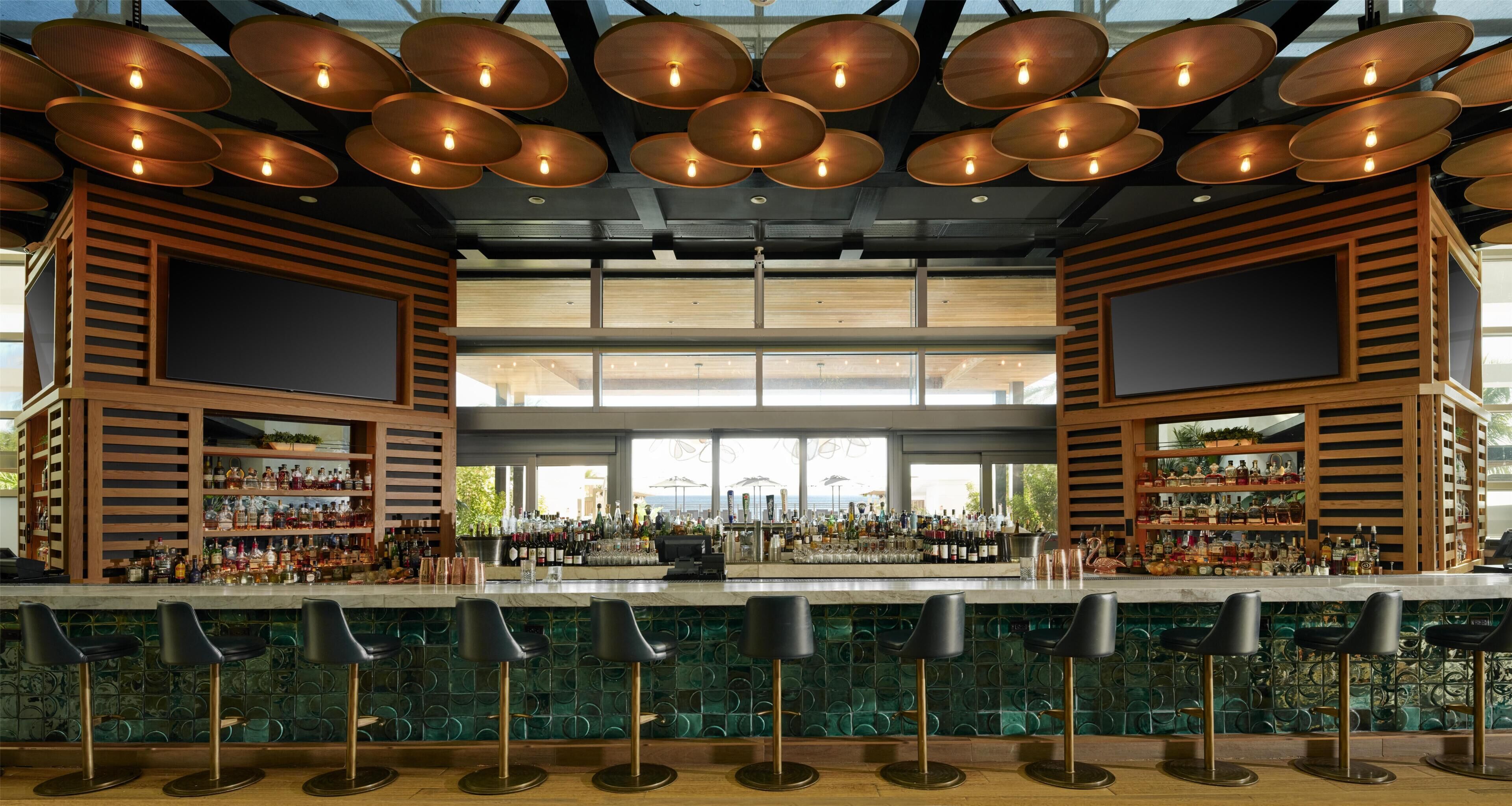 A bar with multiple stools, a variety of bottles, and a large window in the background.