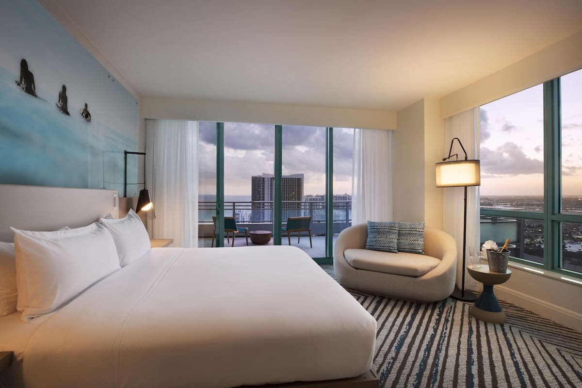 A hotel room with a large bed, a seating area with a chair and a small table, a floor lamp, and a view of the city through large windows.
