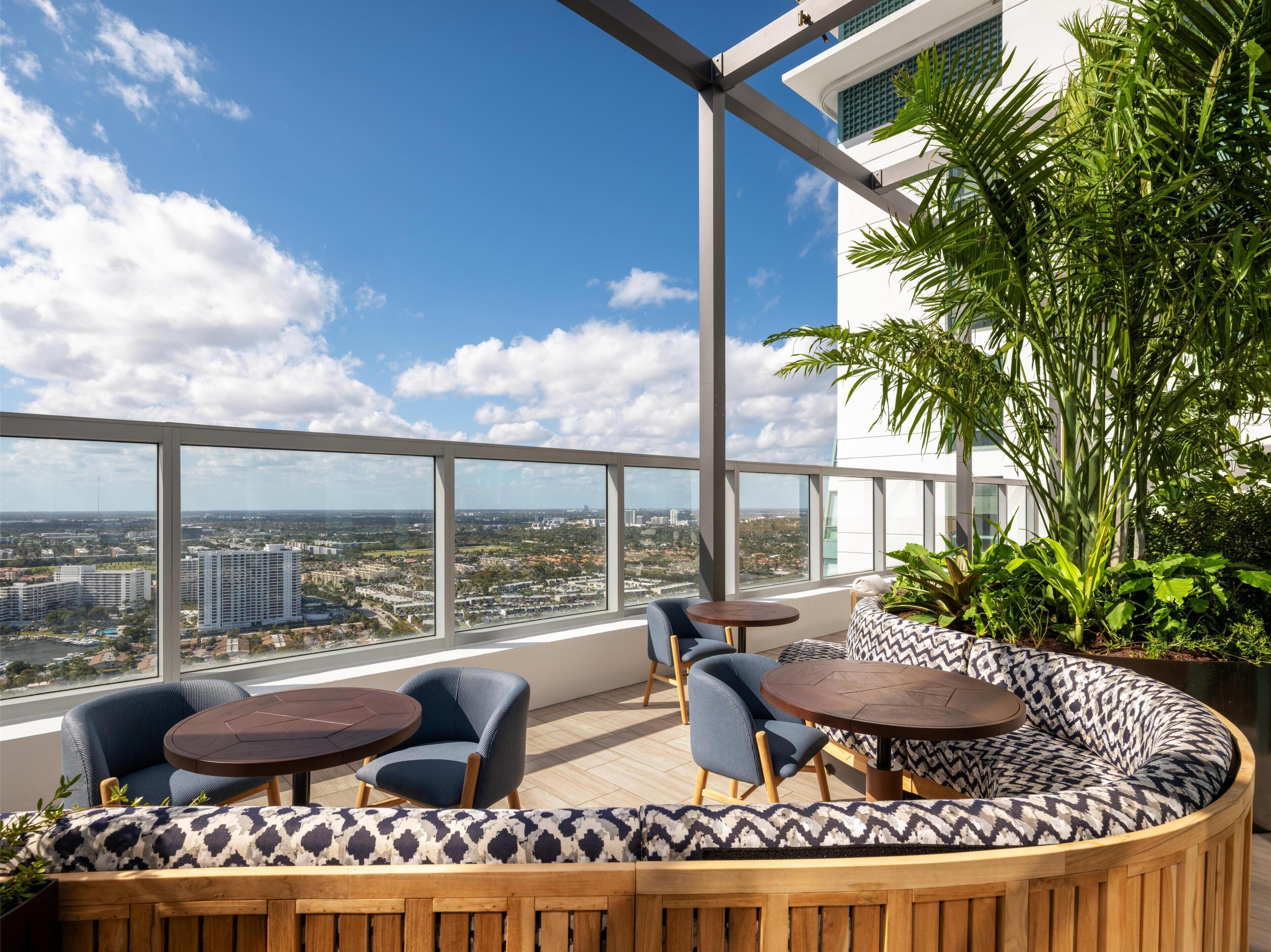 A rooftop terrace with a panoramic city view, furnished seating, and a large potted plant.