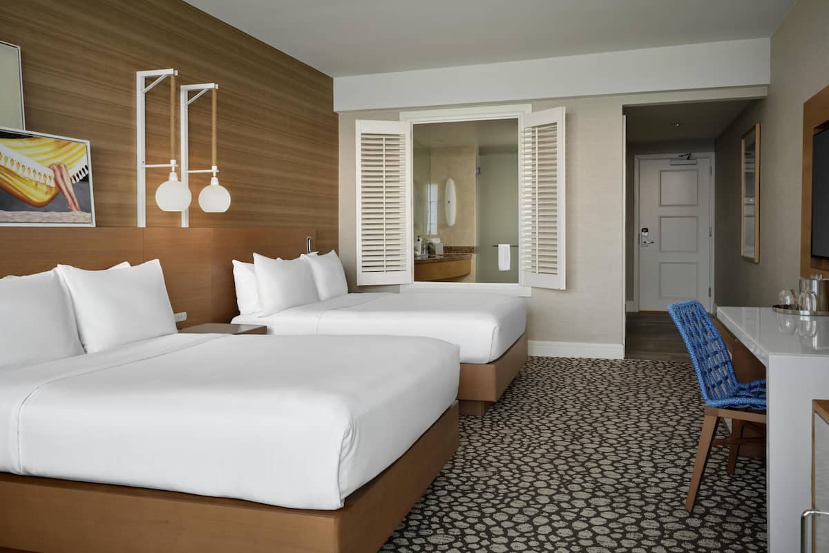 A hotel room with two beds, a desk, a chair, and a bathroom visible through an open door.
