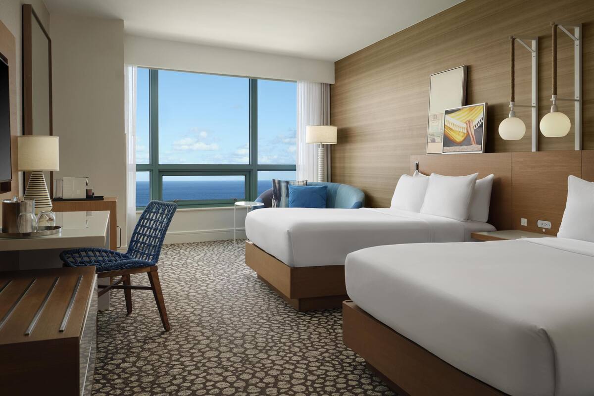 A hotel room with two beds, a desk with a chair, a window with a view of the ocean, and a wall-mounted lamp.