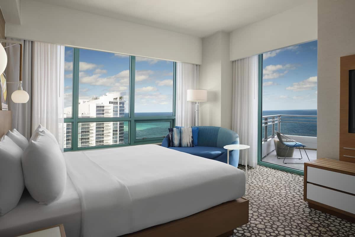 A hotel room with a large bed, a blue sofa, a small table, a lamp, and a view of the ocean.