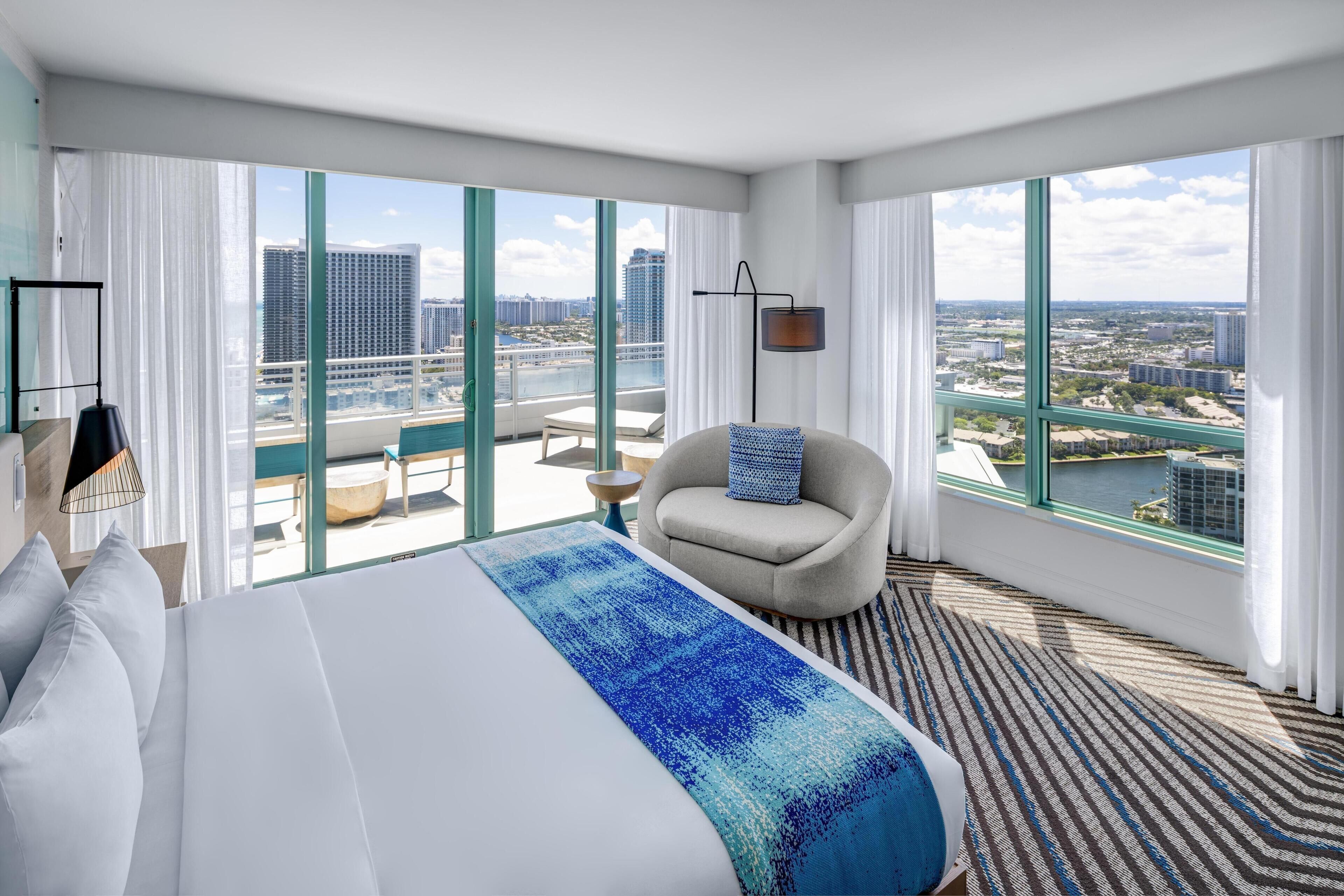 A hotel room with a large bed, a chair, a view of the city, and a balcony.