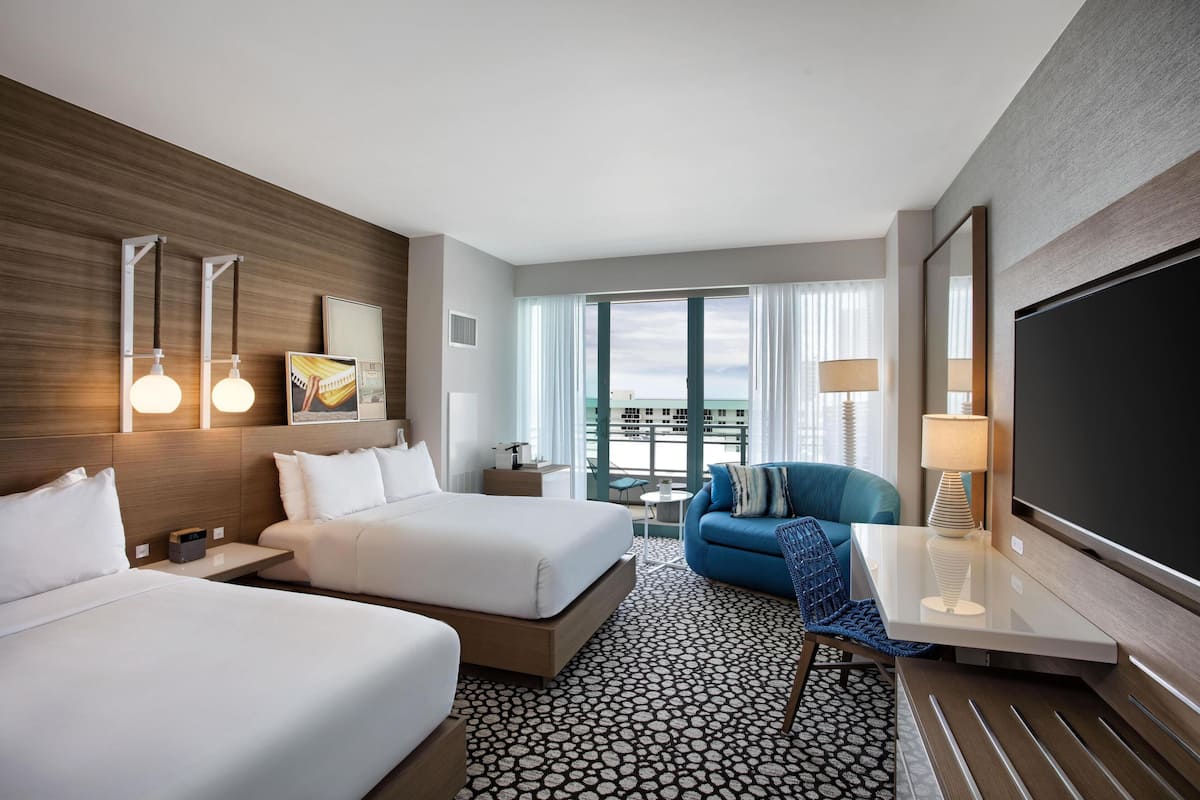 A hotel room with two beds, a TV, a desk with a lamp, a chair, and a view of the city.