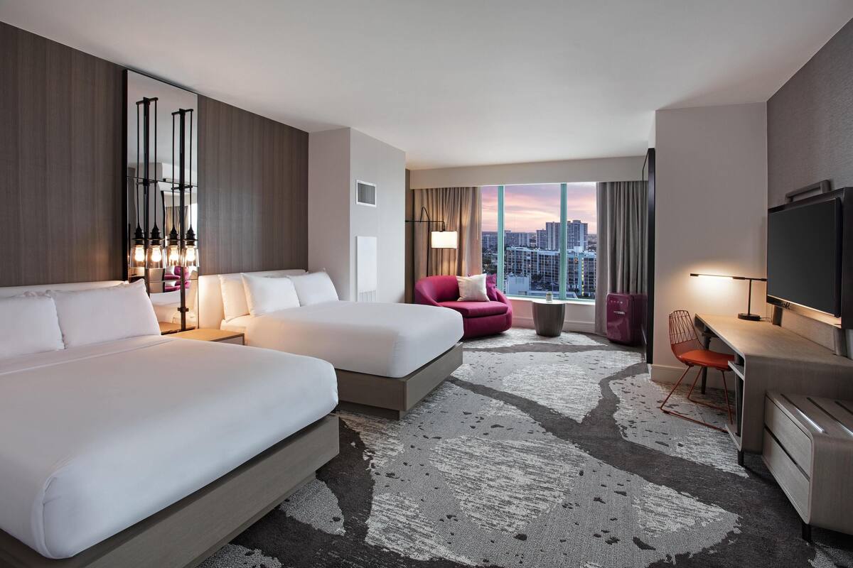 A hotel room with two beds, a desk, a chair, a TV, and a large window with a city view.