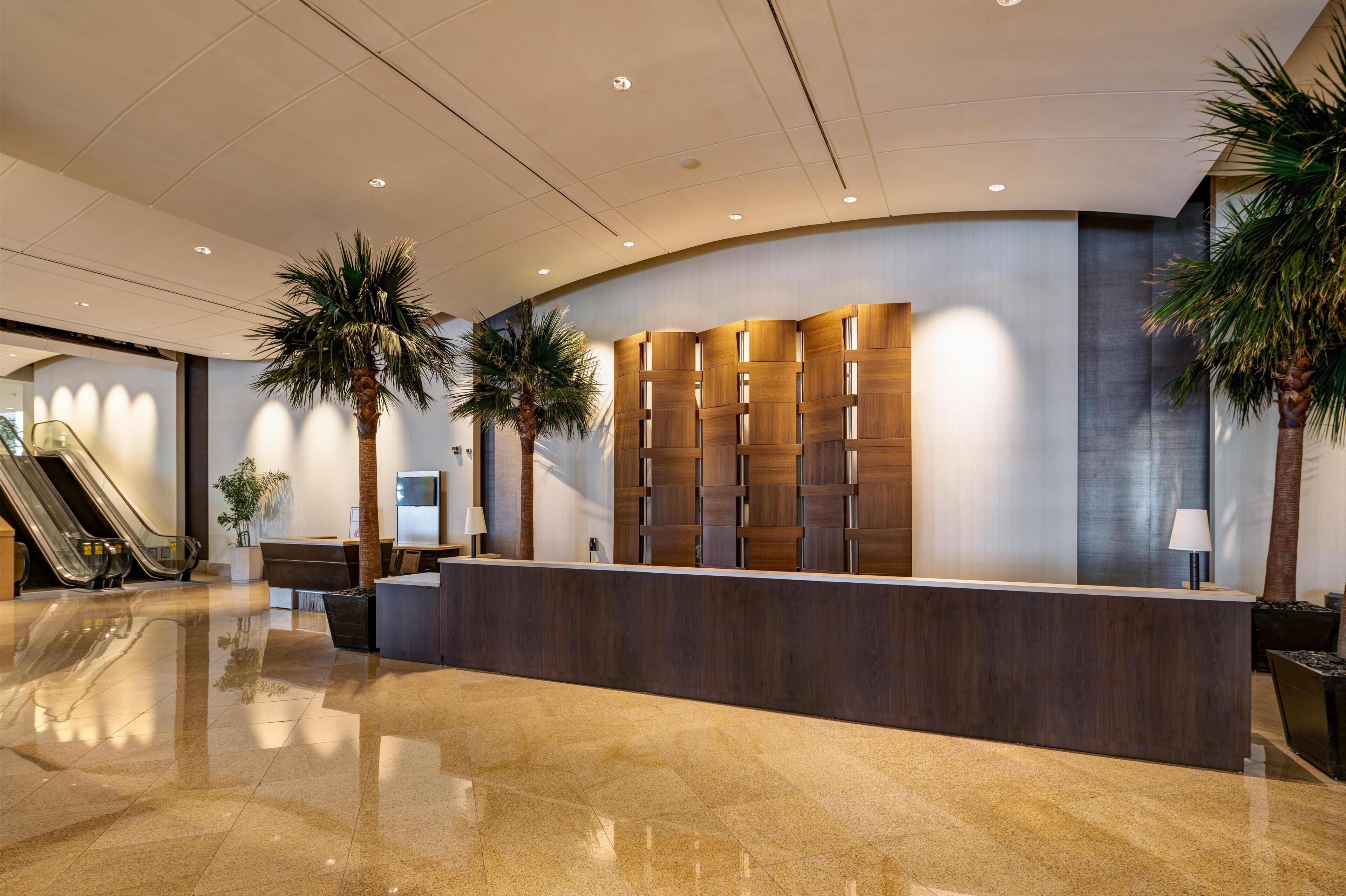 A modern reception area with a curved reception desk, palm trees, and an escalator.