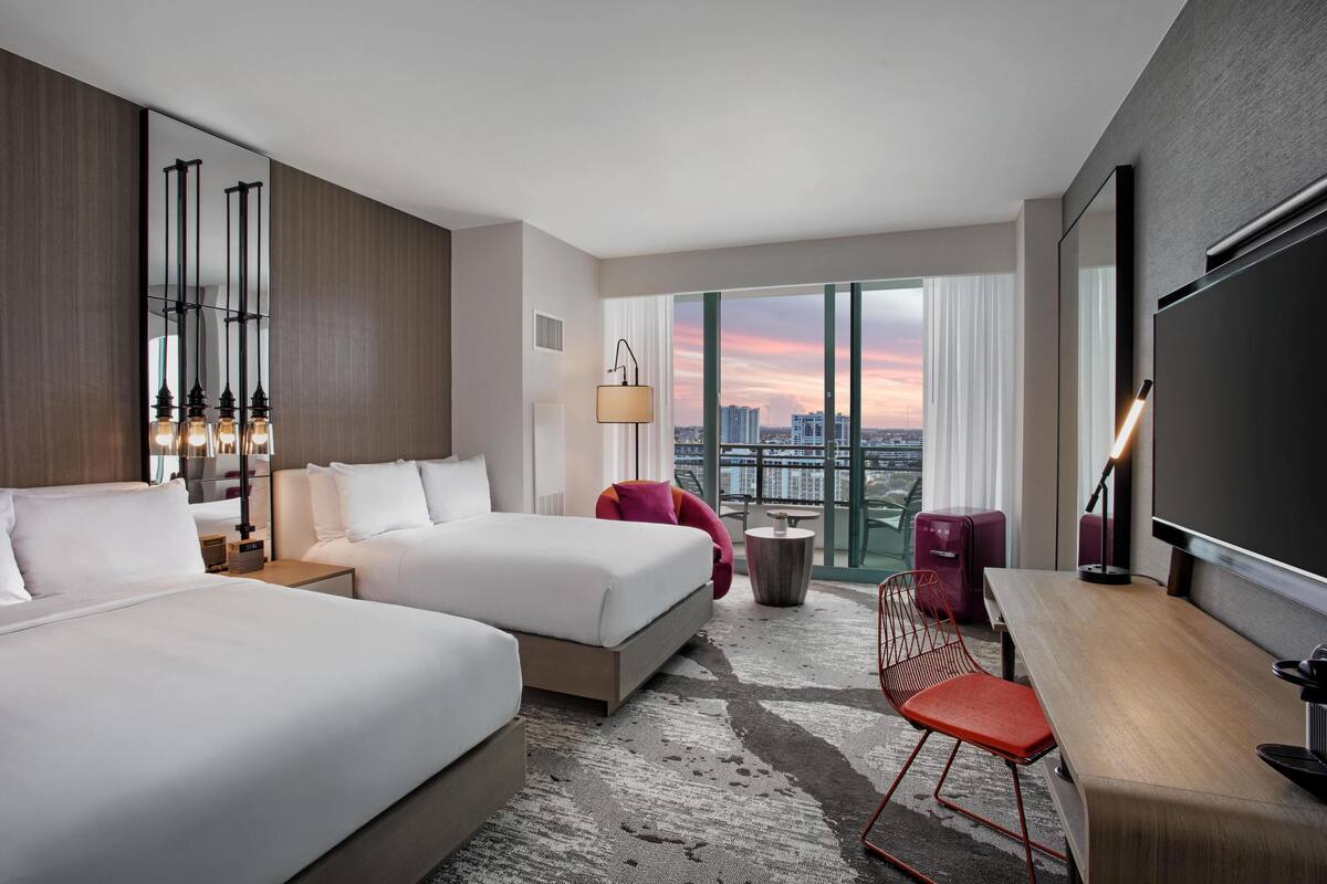 A hotel room with two beds, a desk, a chair, a TV, a lamp, and a view of the city.