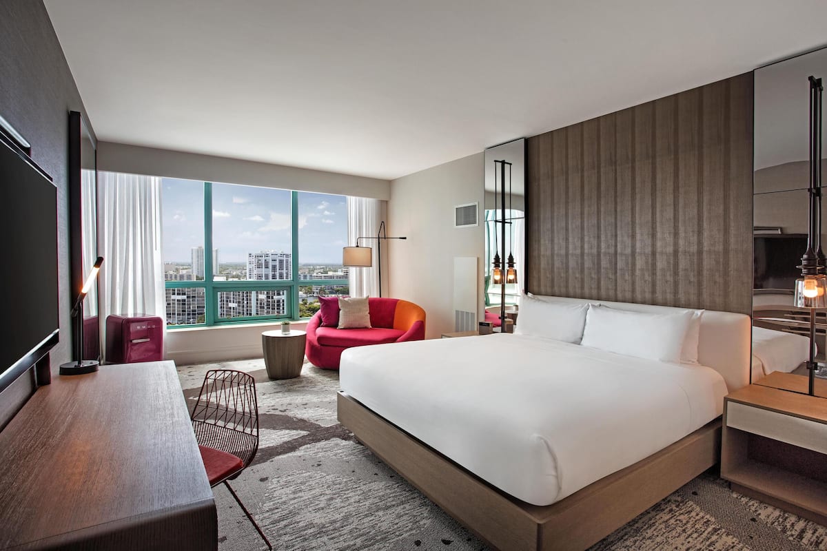 A hotel room with a large bed, a desk, a red chair, a small table, a mirror, and a view of the city.