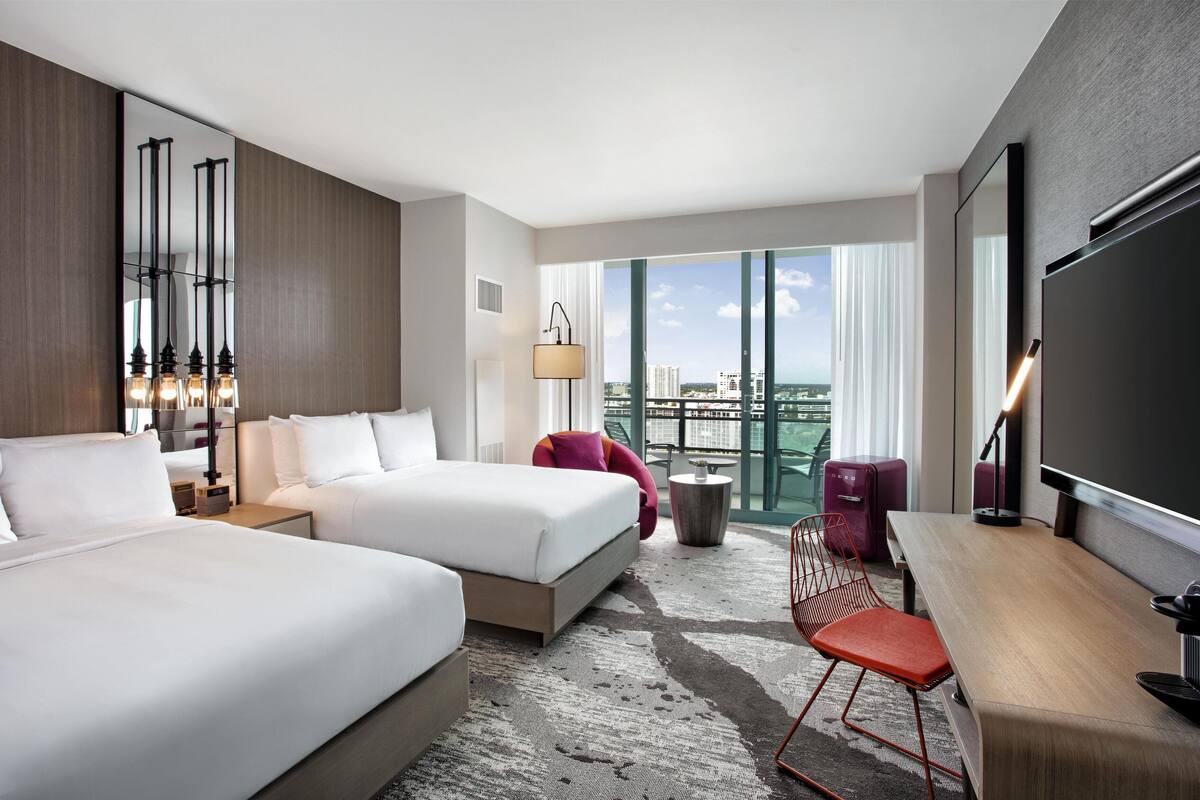 A hotel room with two beds, a desk, a chair, a TV, and a balcony with a city view.